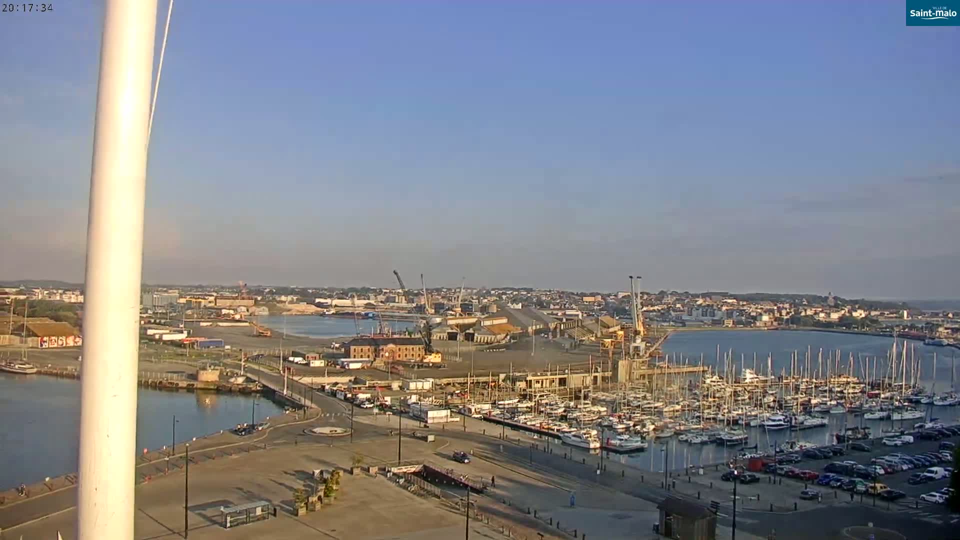 A harbor filled with boats is shown under a clear, sunny sky, with construction taking place near a waterfront city.