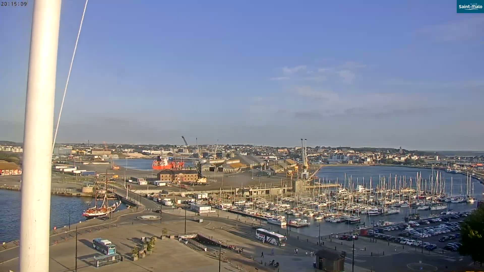 A harbor with numerous sailboats and yachts moored, a large cargo ship, and industrial buildings under a clear, sunny sky.