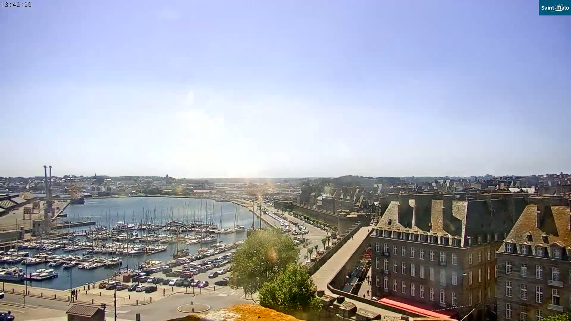 A sunny day overlooking a harbor filled with sailboats, a parking lot, and a town with stone buildings.