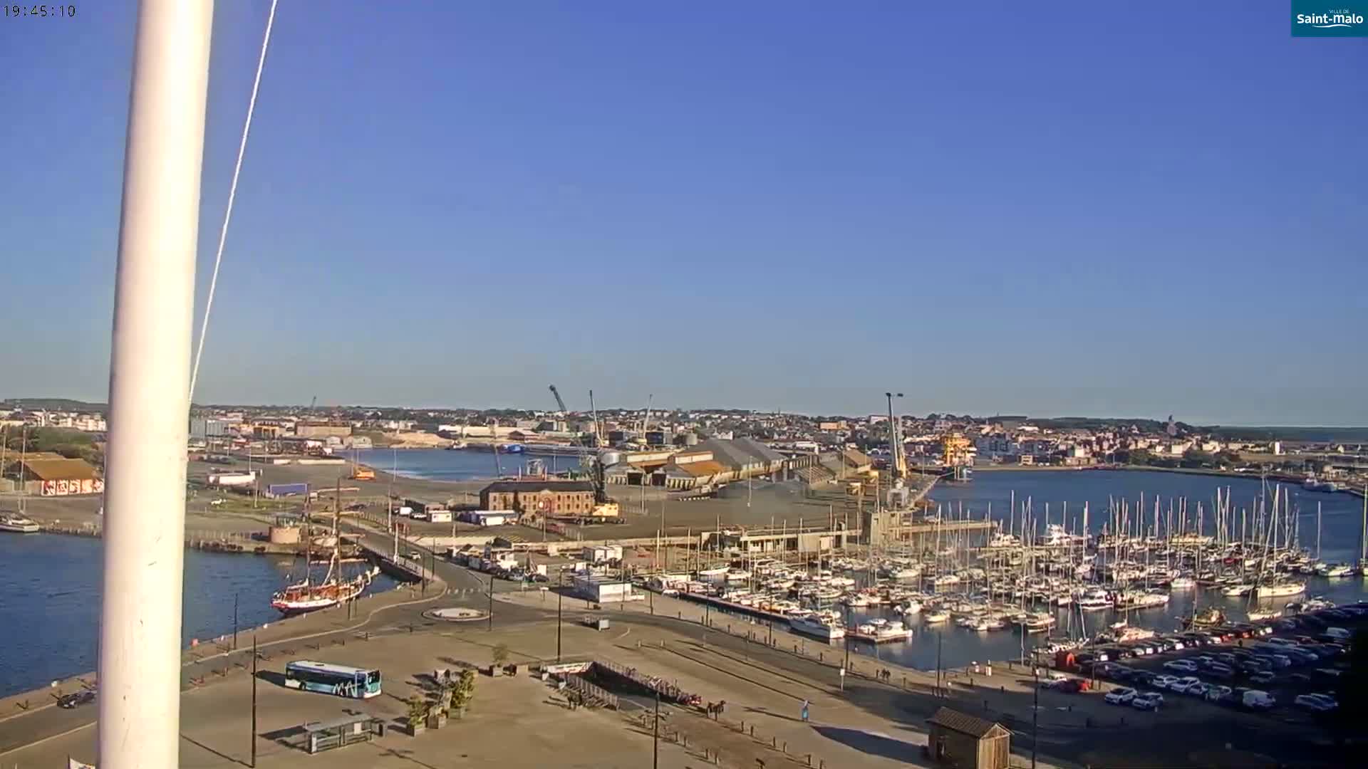 A harbor with numerous docked sailboats and a small city in the background under clear, sunny skies.