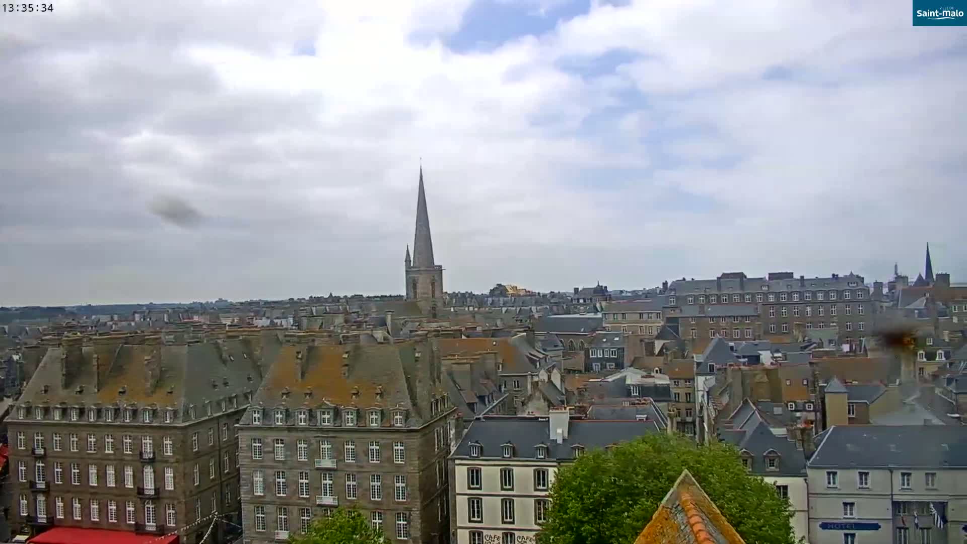 A partly cloudy sky overlooks a city of grey-roofed buildings, with a tall church spire visible in the mid-ground.