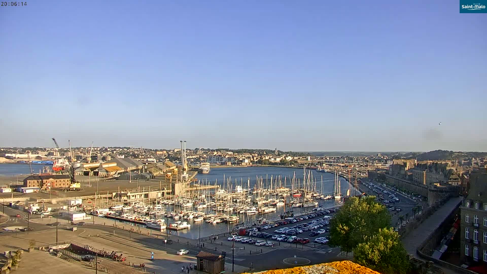 A harbor filled with sailboats and cars is situated next to a walled city under a clear blue sky.