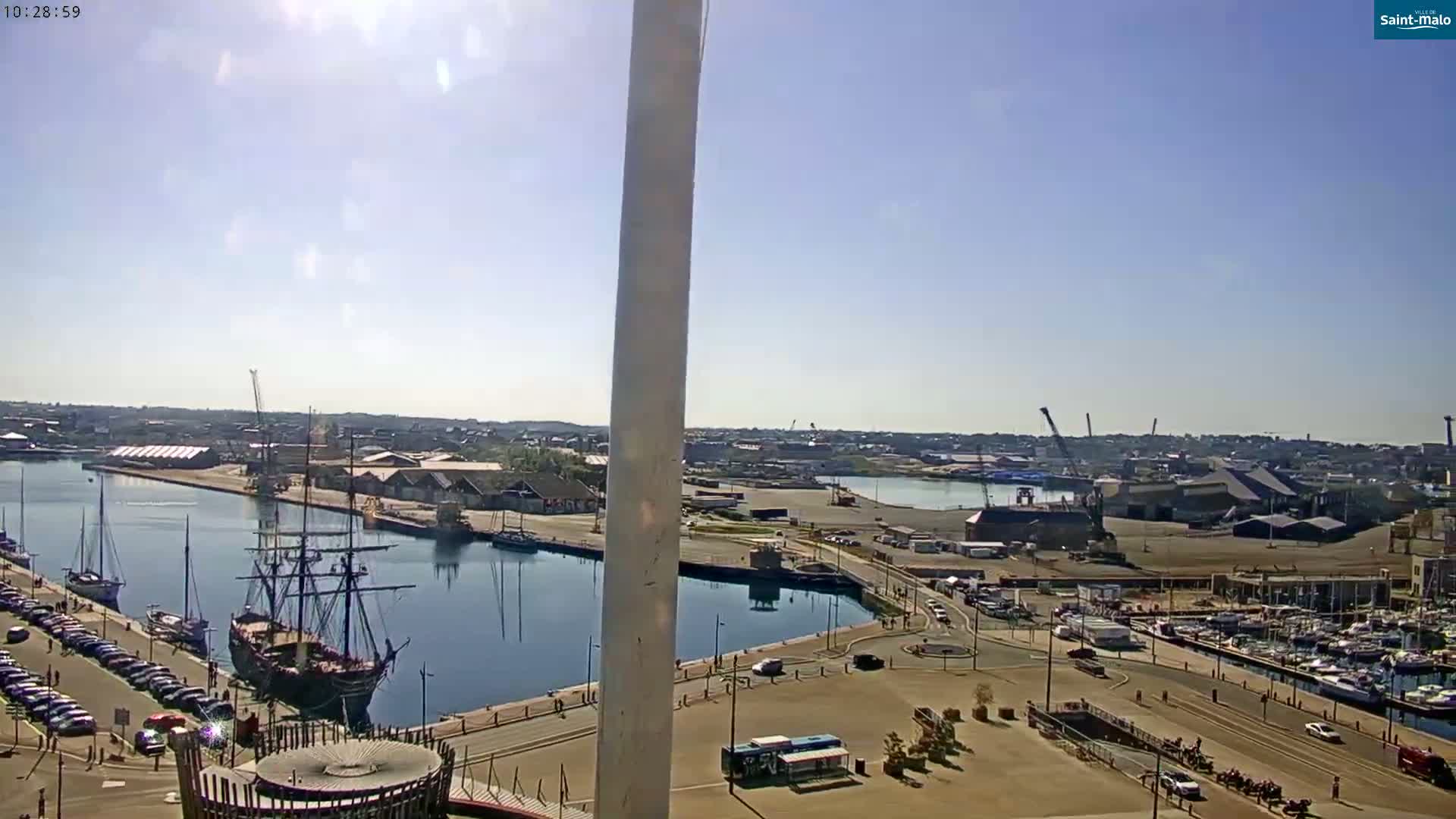 A sunny day overlooking a harbor with a large sailing ship docked, numerous smaller boats moored nearby, and a city in the background.