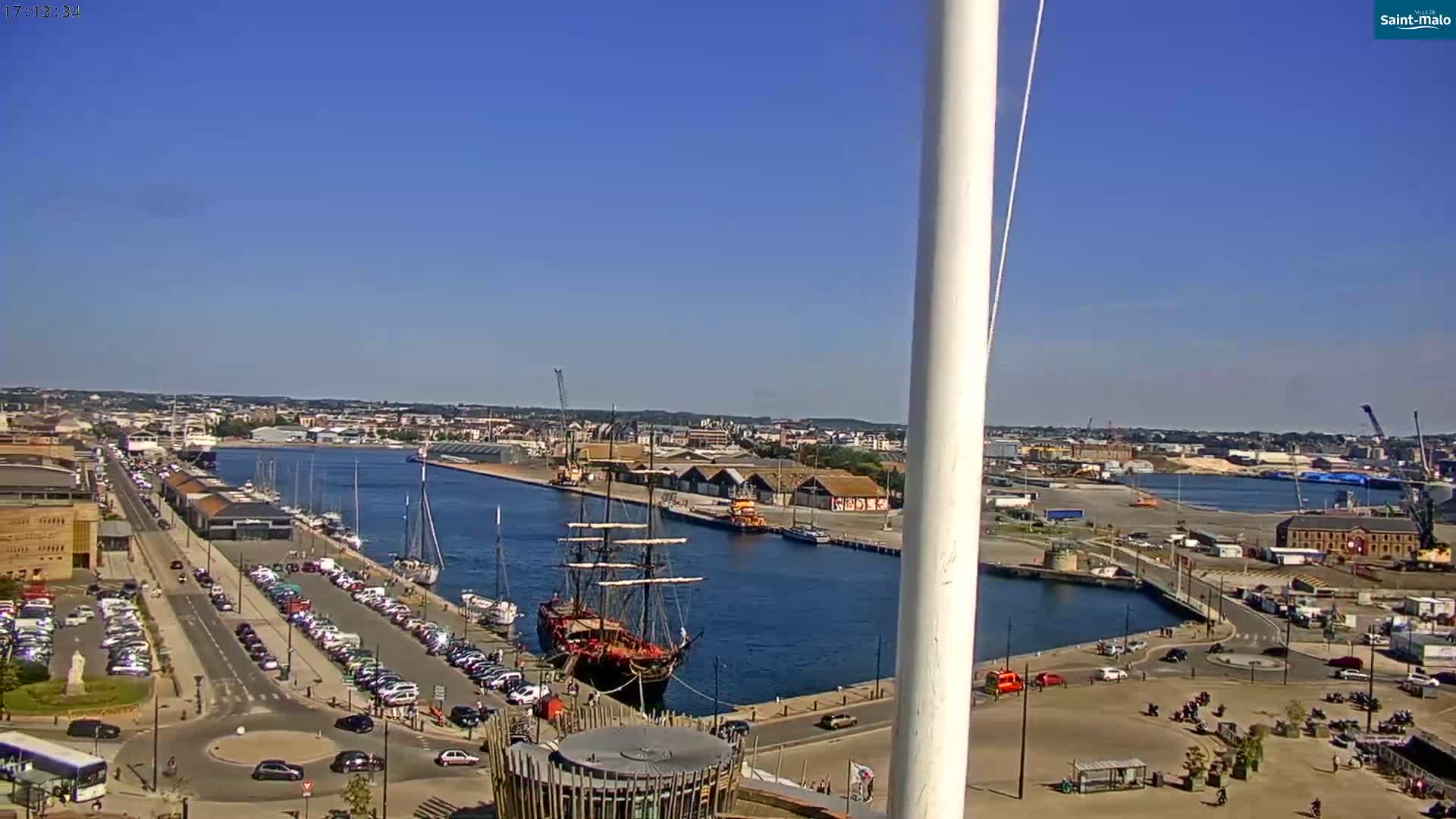 A large sailing ship is docked in a harbor on a sunny day, with numerous other boats and cars visible along the waterfront.