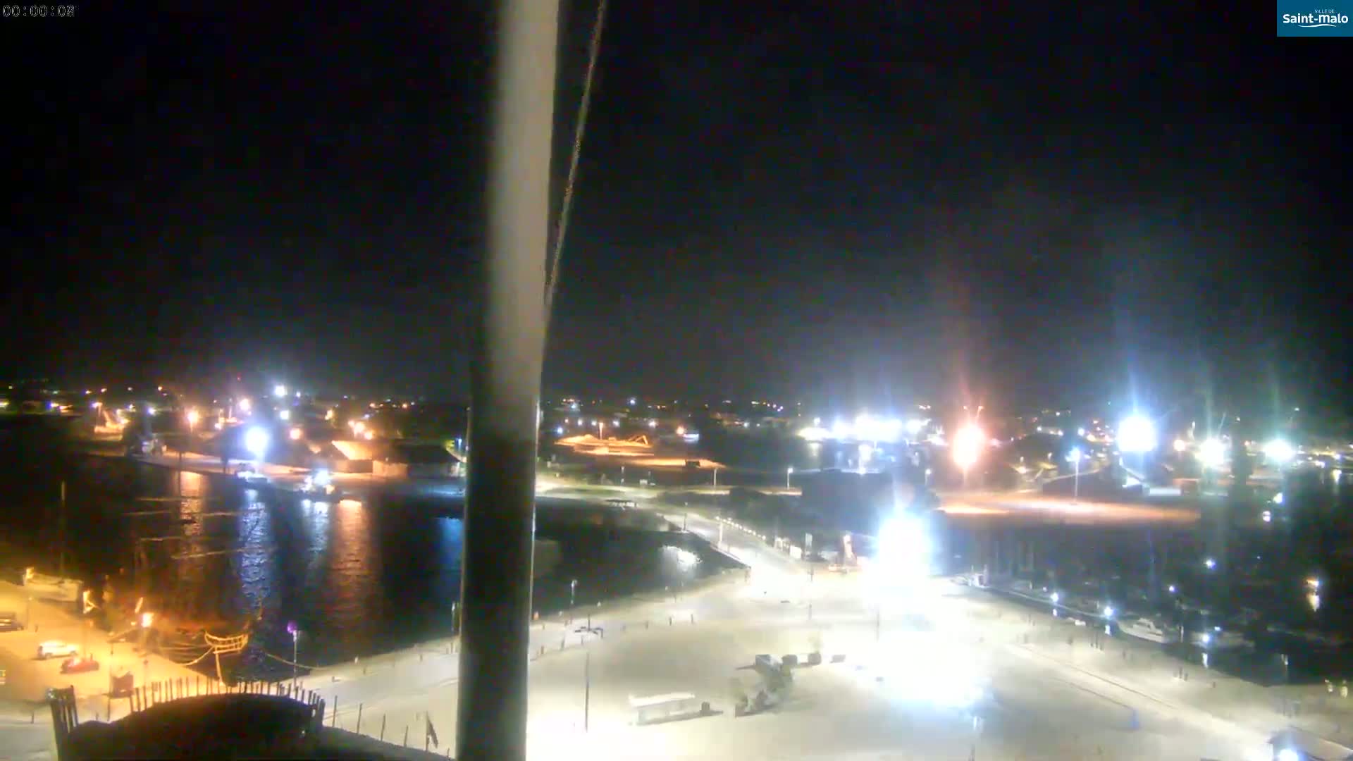 A nighttime view of a harbor with brightly lit docks and several boats, under a clear, dark sky.