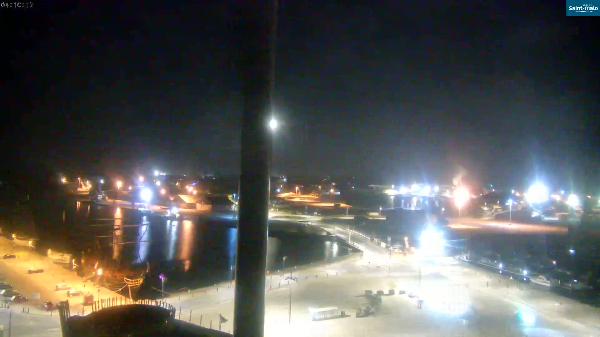 A nighttime, high-angle view shows a harbor with a large ship, brightly lit buildings, and a mostly empty parking lot.