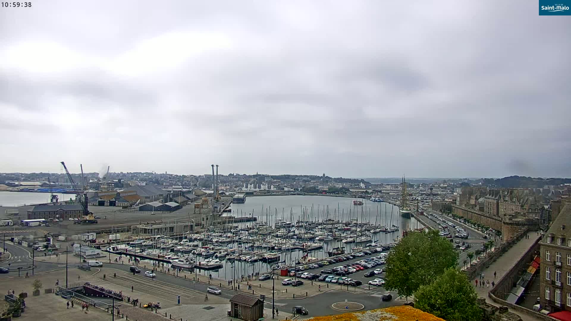 A harbor filled with numerous sailboats and yachts under an overcast sky, adjacent to a city with buildings and walls.