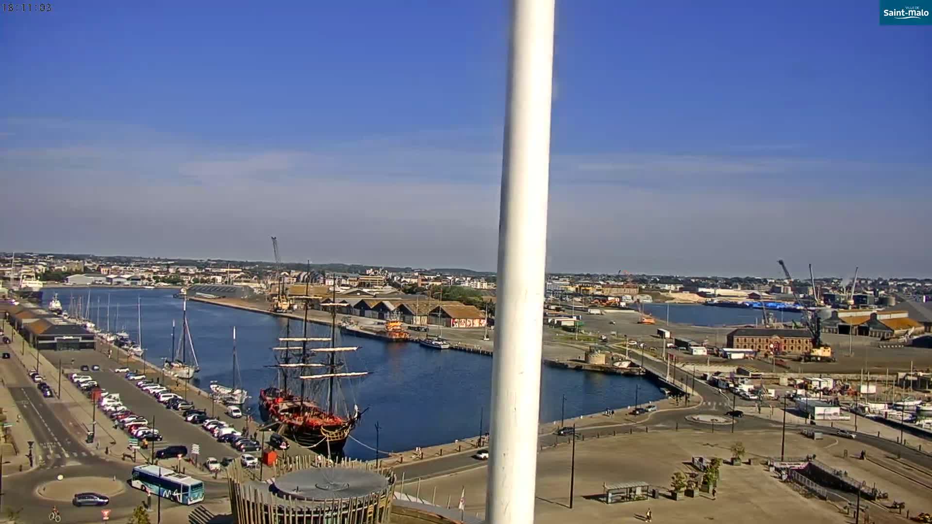 A large sailing ship is docked in a harbor on a sunny day, with numerous other boats and cars visible, and a city in the background.