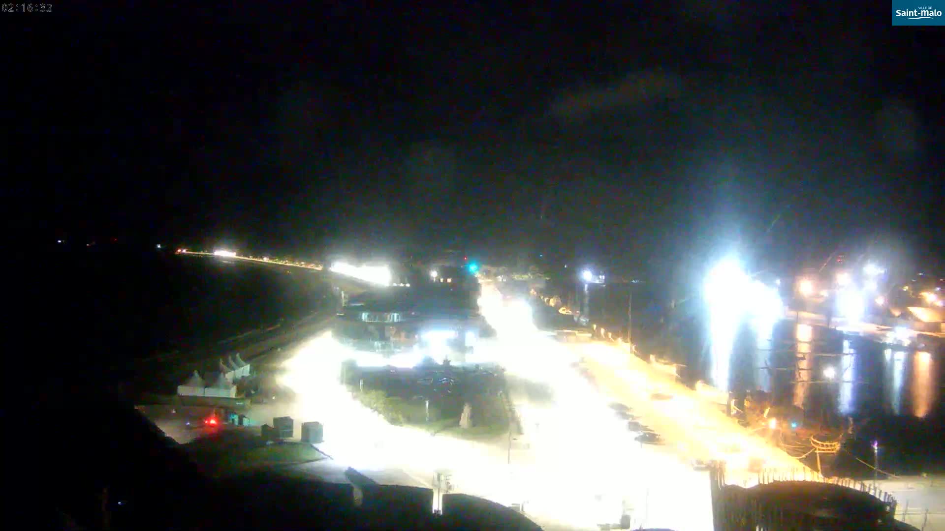 A nighttime, high-angle view shows brightly lit roads and buildings near a body of water, where a ship is docked.