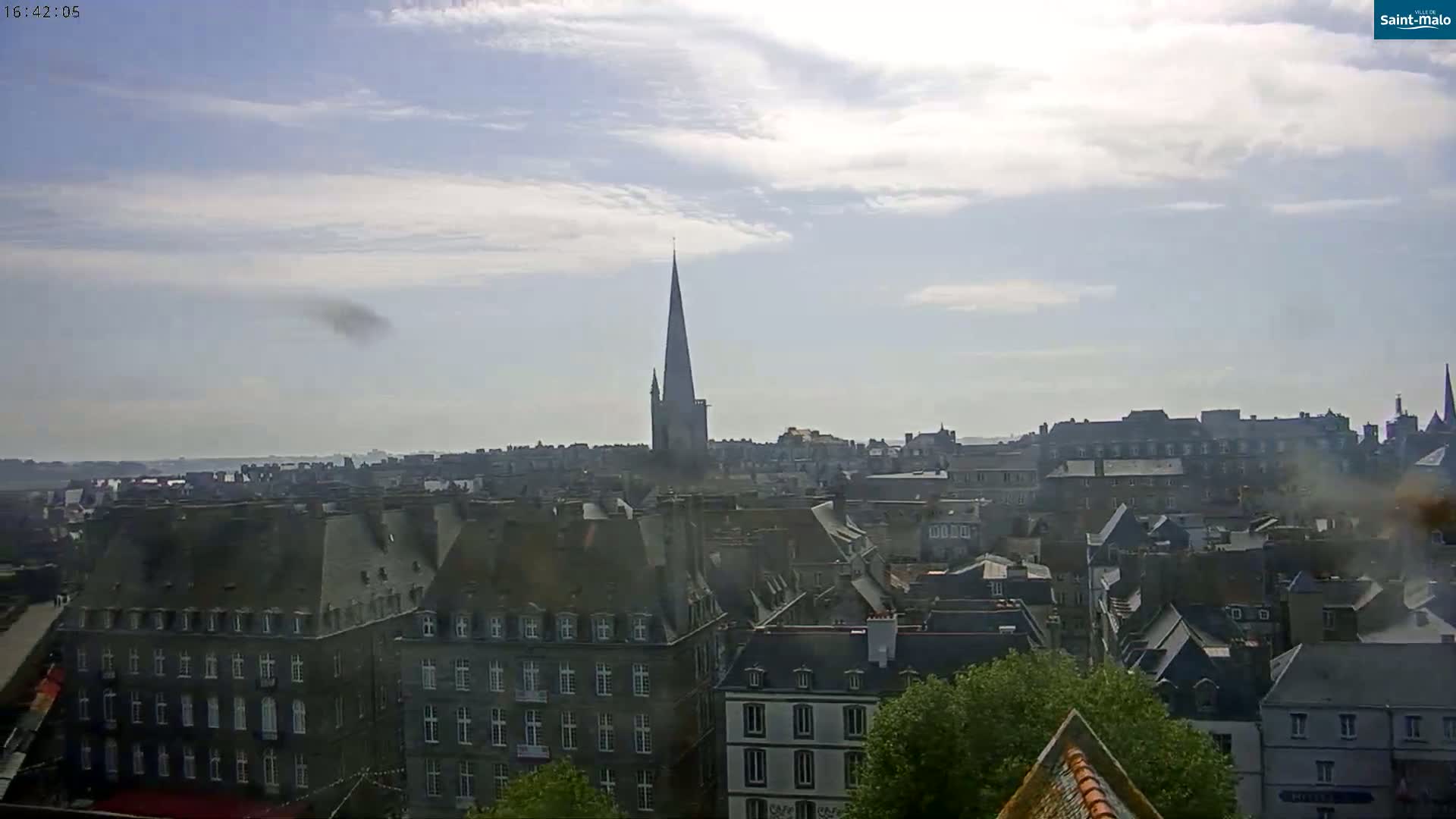 A partly cloudy day overlooks a city with many grey-roofed buildings and a tall, pointed church spire.
