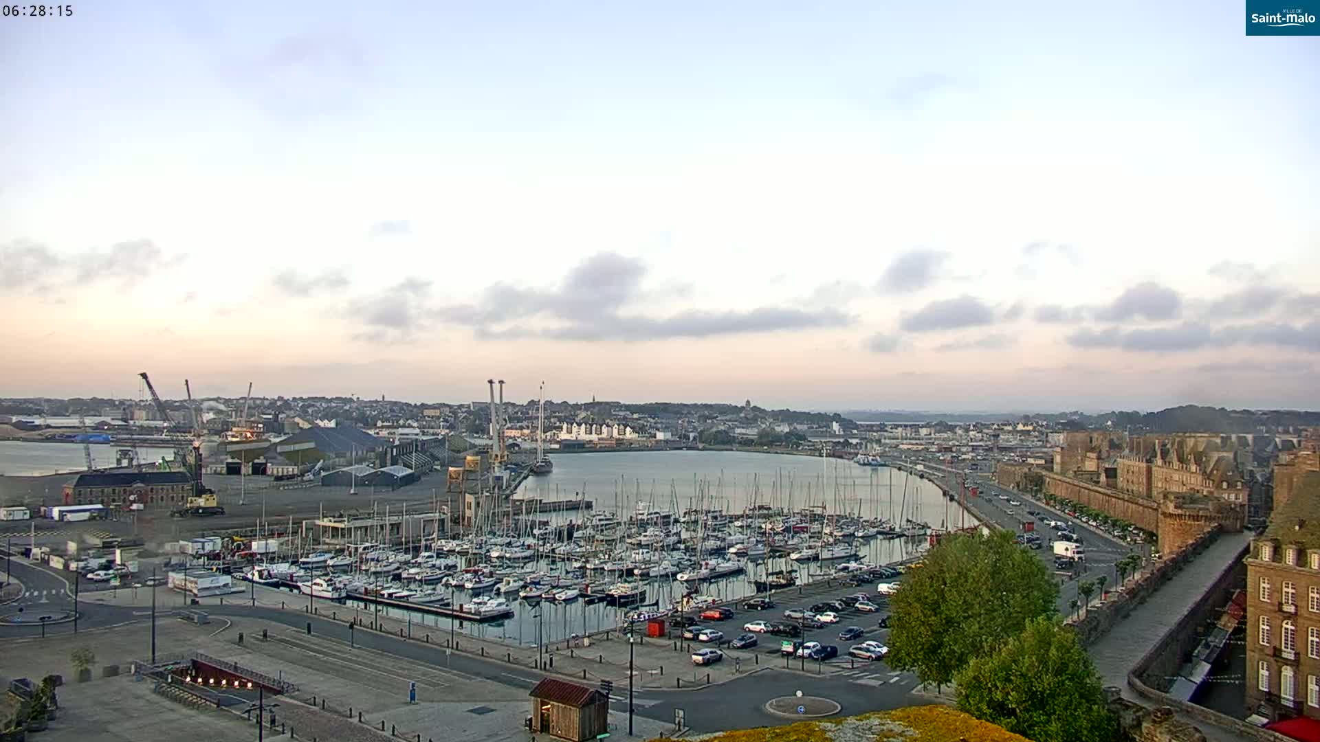 A harbor filled with sailboats is shown at sunrise under a mostly clear sky, next to a stone wall and buildings of a town.