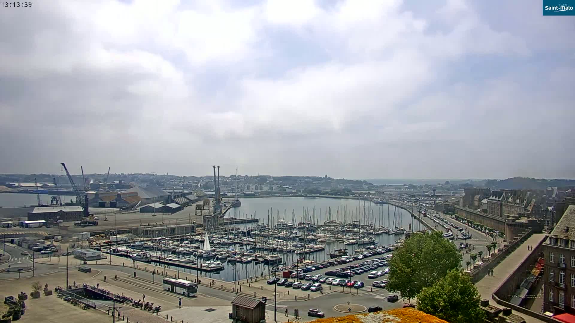 A partly cloudy day overlooks a harbor filled with many sailboats and a city with buildings and parked cars along the water.