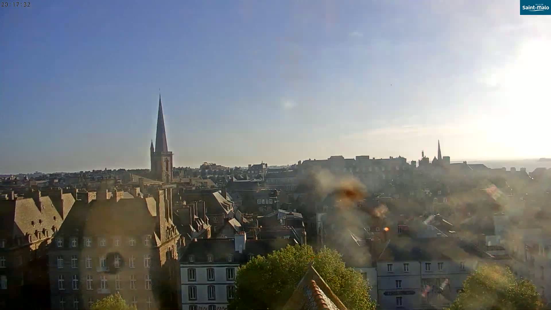 A hazy, sunny view of a city skyline with a tall church steeple and many rooftops, seen from an elevated perspective.
