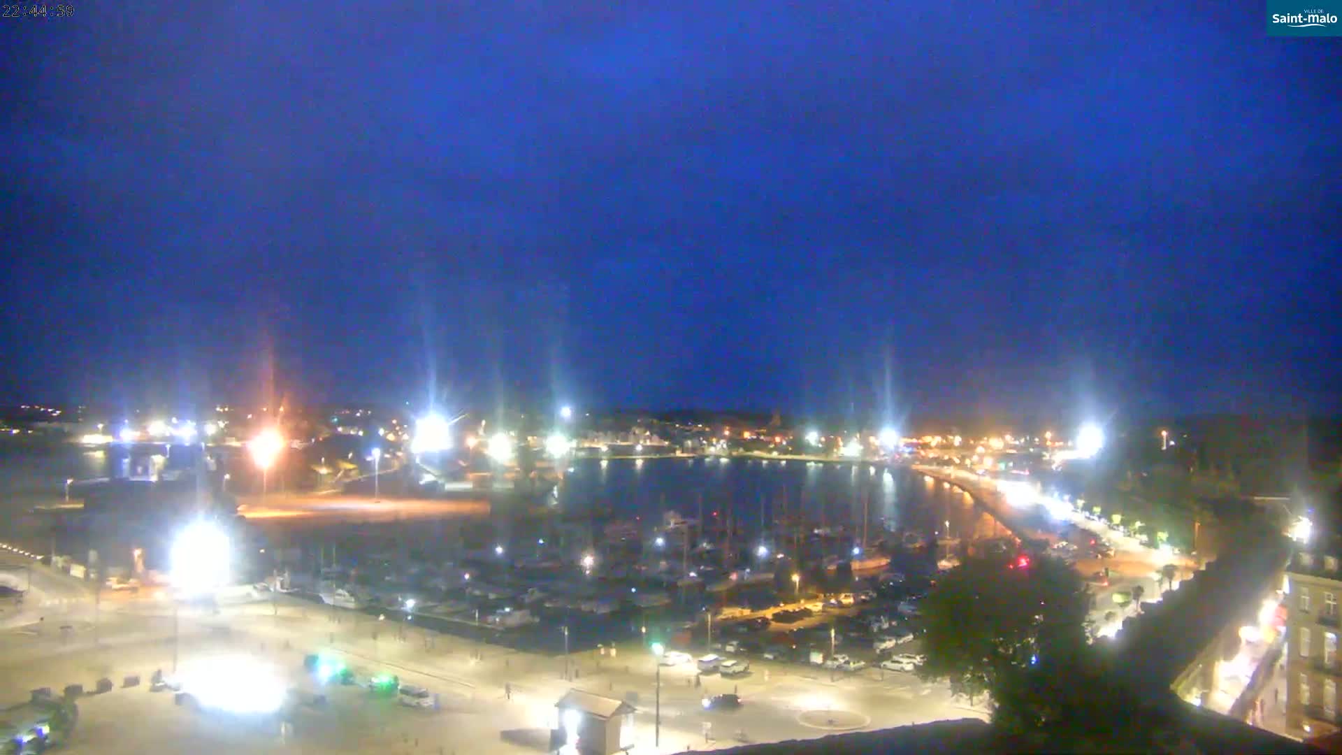 A nighttime view of a harbor filled with boats, surrounded by illuminated buildings and streets under a clear, dark-blue sky.