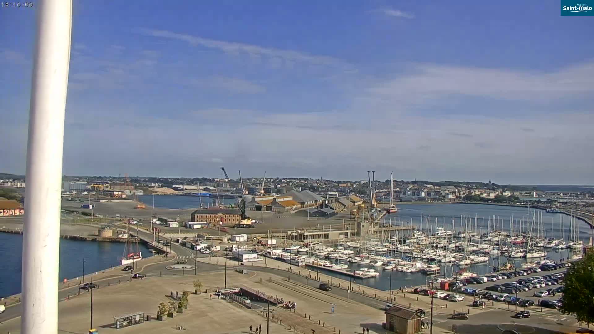 A harbor filled with numerous sailboats and yachts under a mostly sunny sky is seen from an elevated vantage point, with a city and industrial area in the background.