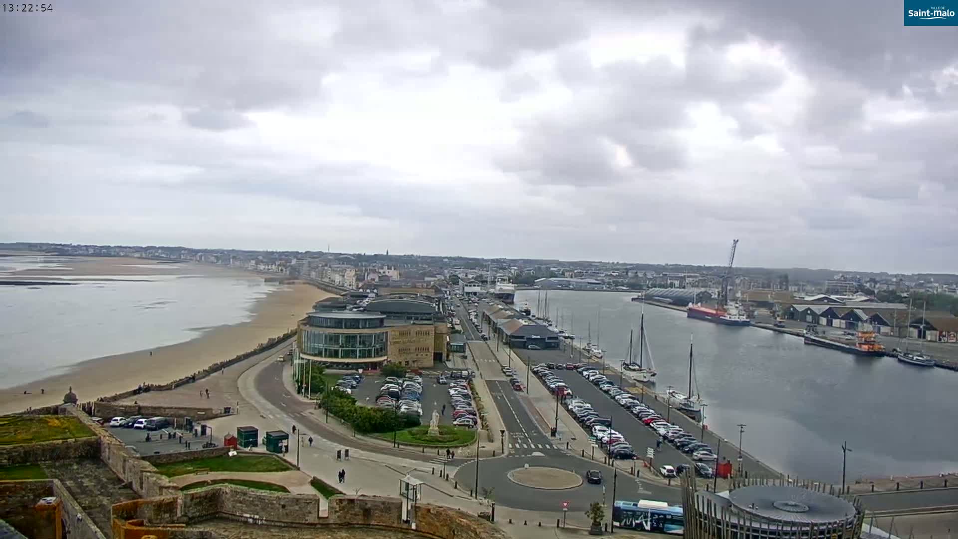 An elevated view shows a coastal city on an overcast day, with a harbor filled with boats and ships, a sandy beach, and parking lots near modern buildings.