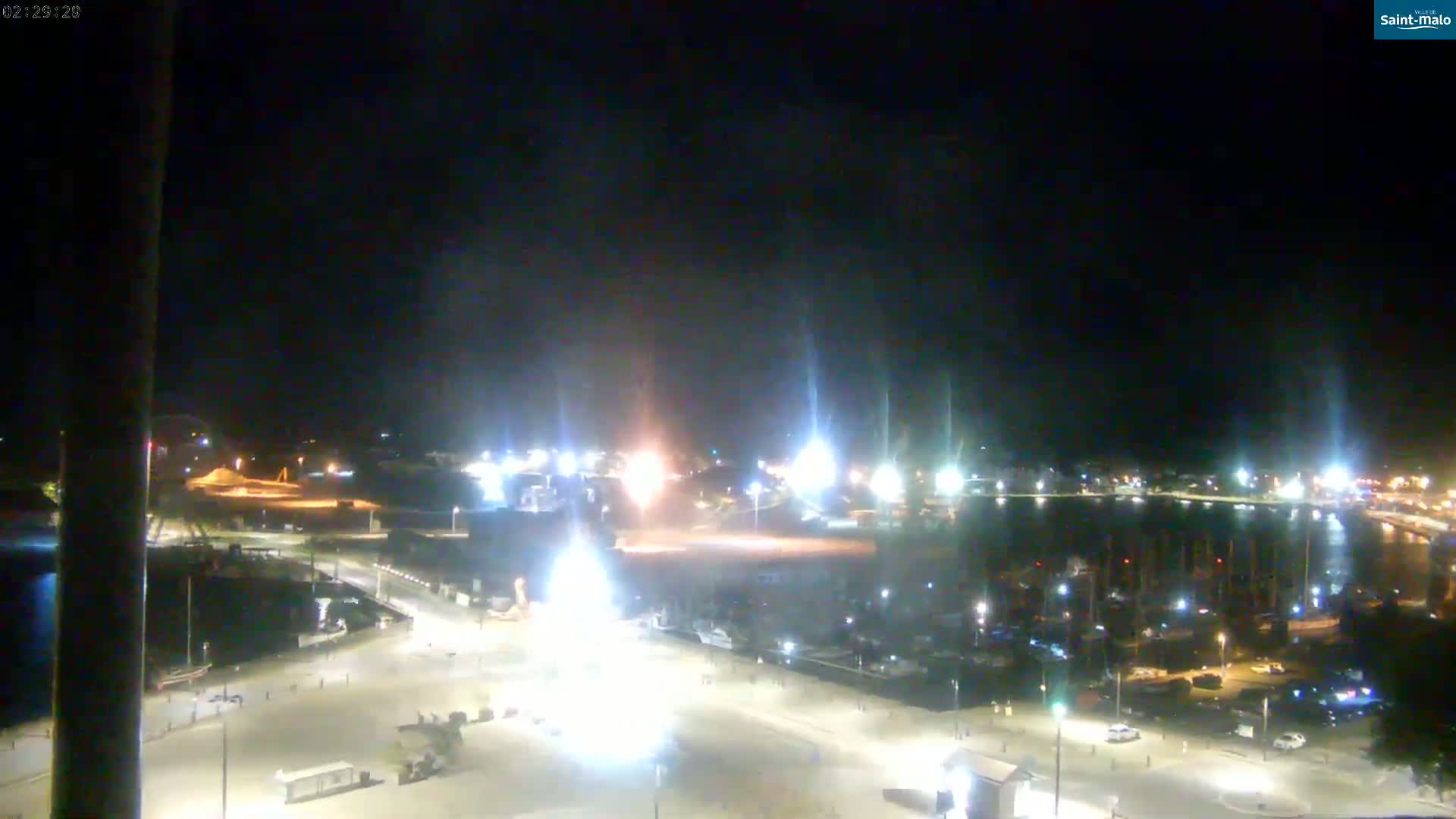 A nighttime, high-angle view of a harbor with many boats docked, surrounded by brightly lit streets and buildings.