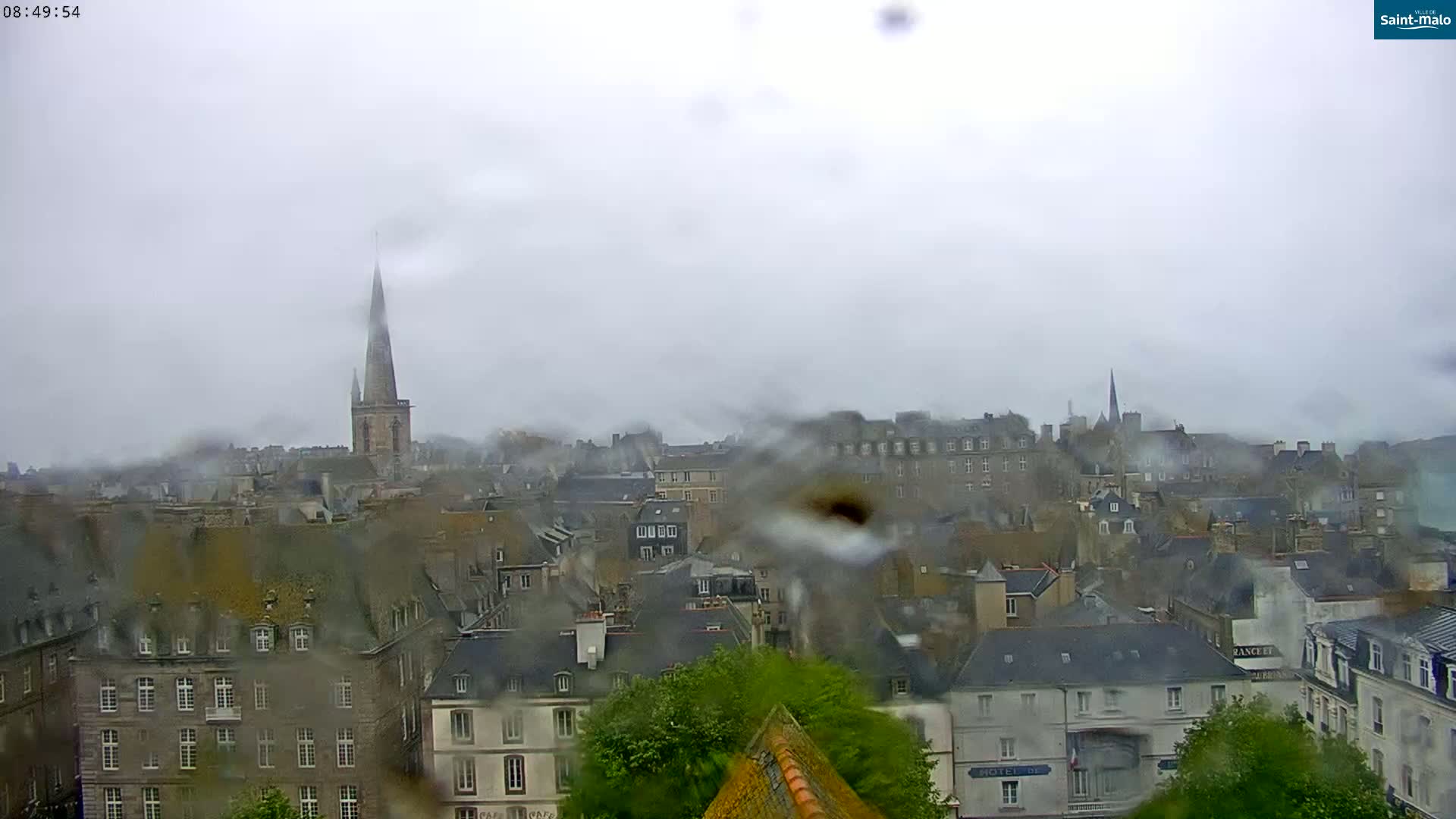 A rain-streaked view of a city with numerous grey buildings and a tall church spire in the distance under an overcast sky.