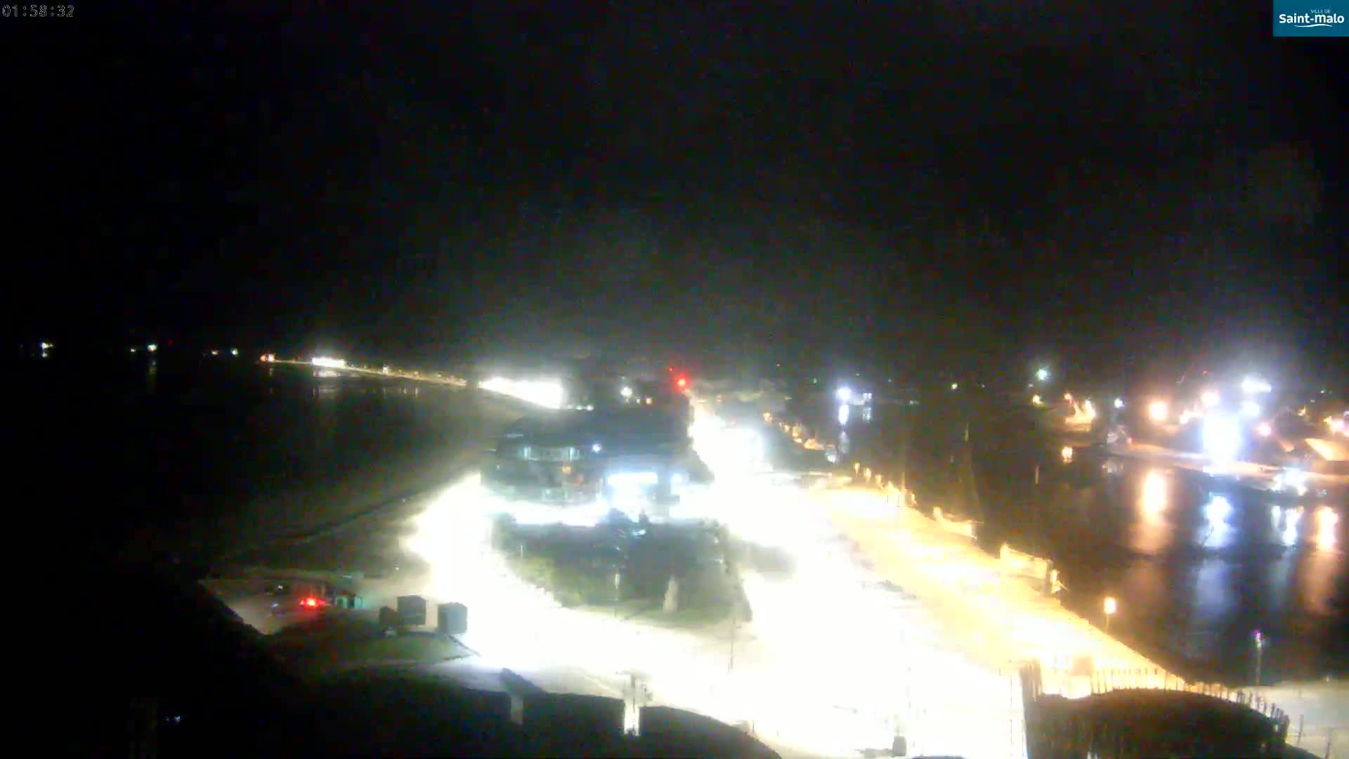 A nighttime, high-angle view shows a waterfront area with roads illuminated by streetlights, buildings, and a dark body of water.