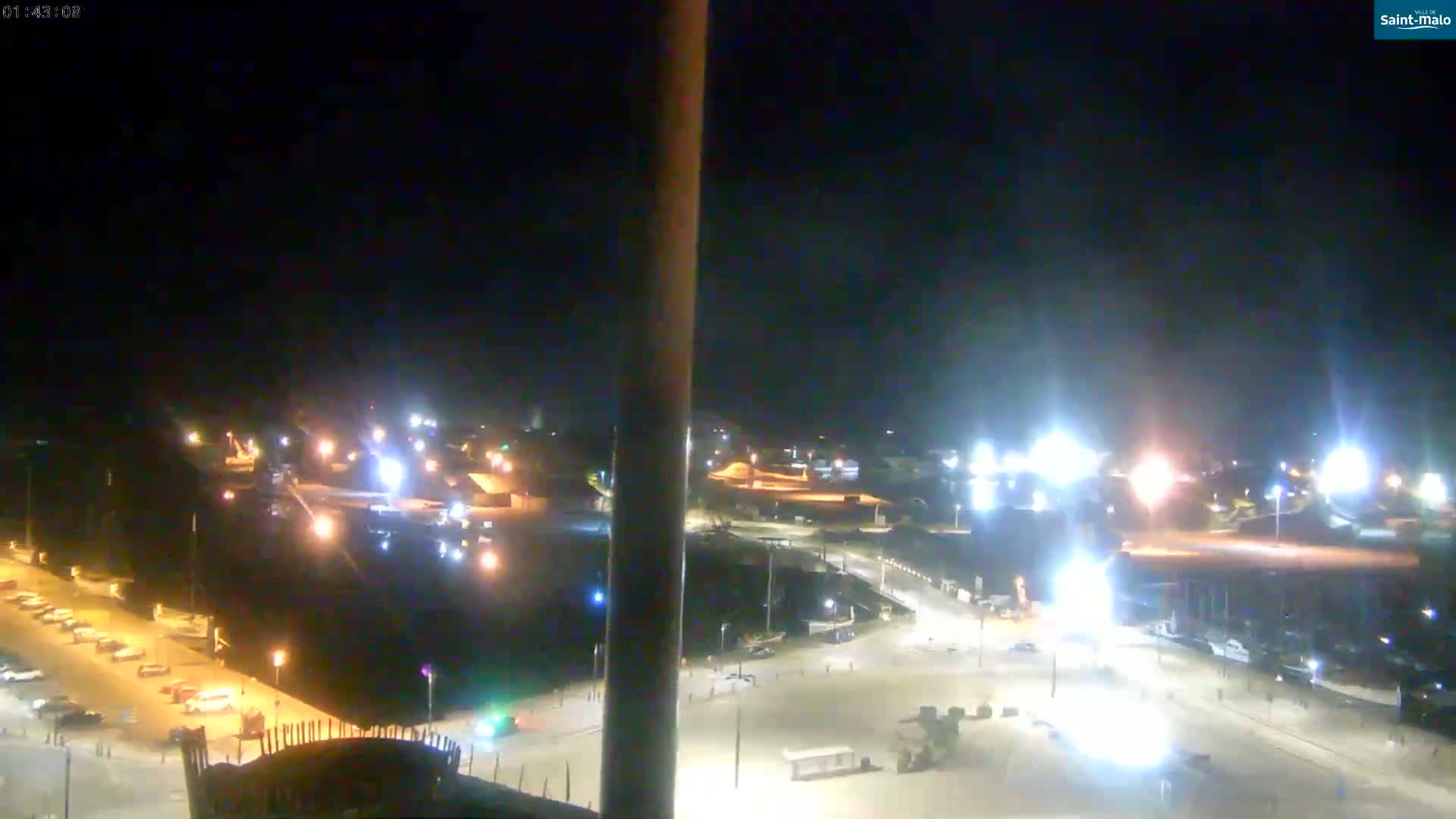 A nighttime aerial view shows a brightly lit harbor area with parked cars, buildings, and various pieces of machinery.