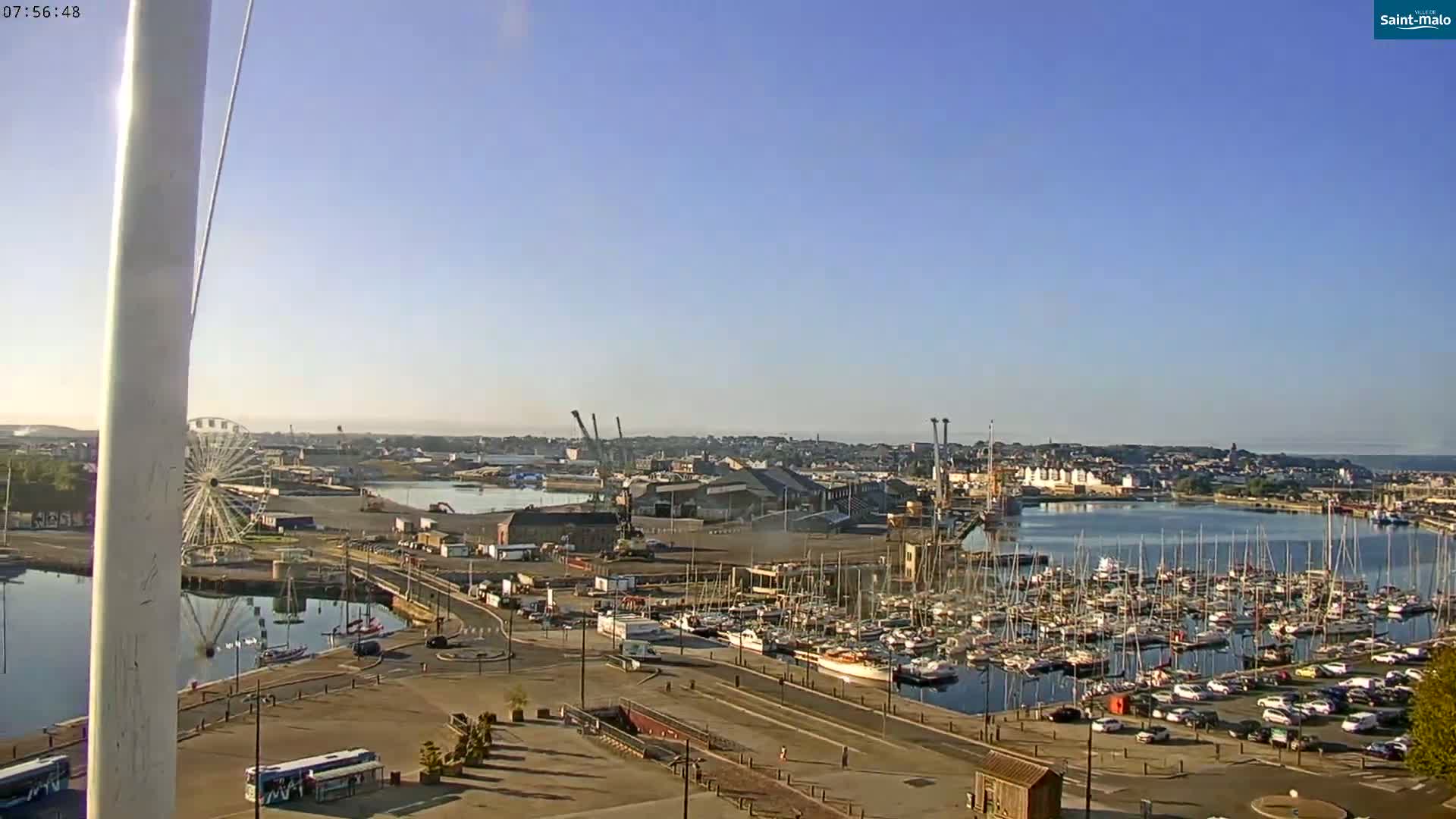 A harbor filled with numerous boats and yachts under a clear, sunny sky, with a Ferris wheel and industrial structures visible in the background.