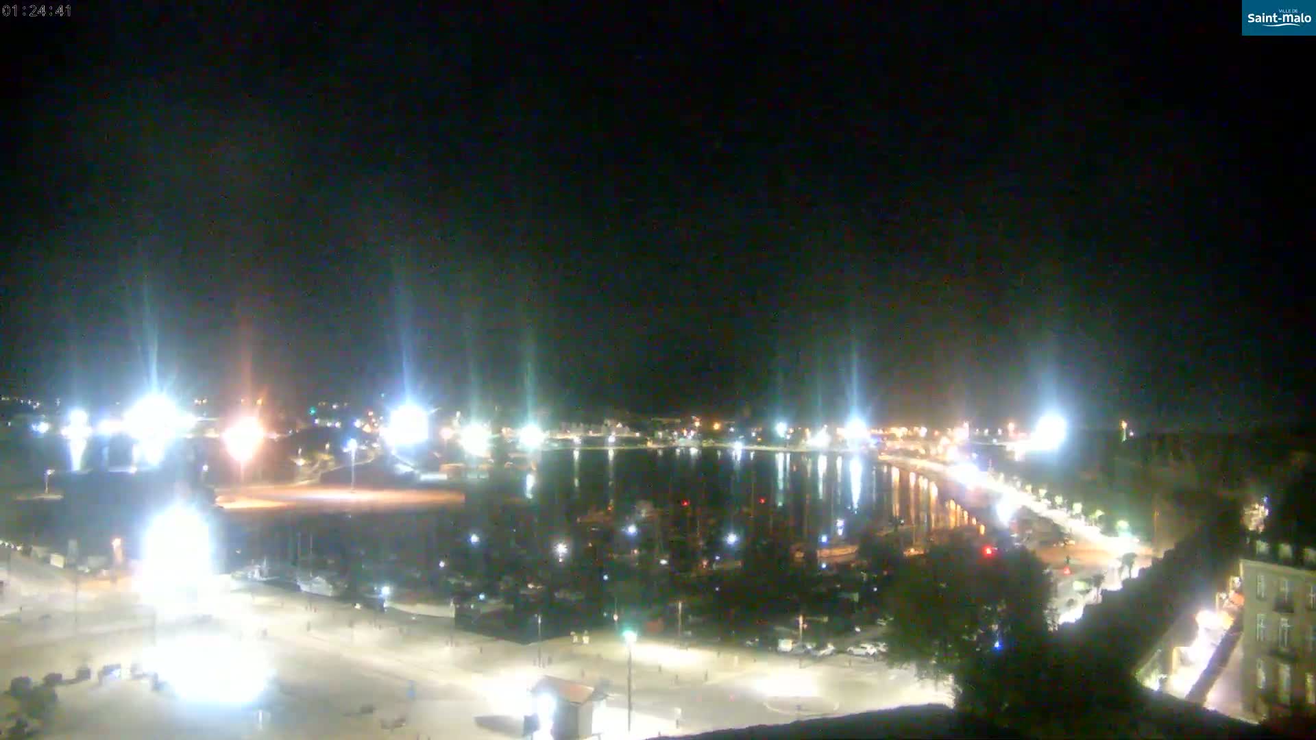 A nighttime aerial view of a harbor filled with boats, illuminated by bright lights along the waterfront under a clear, dark sky.