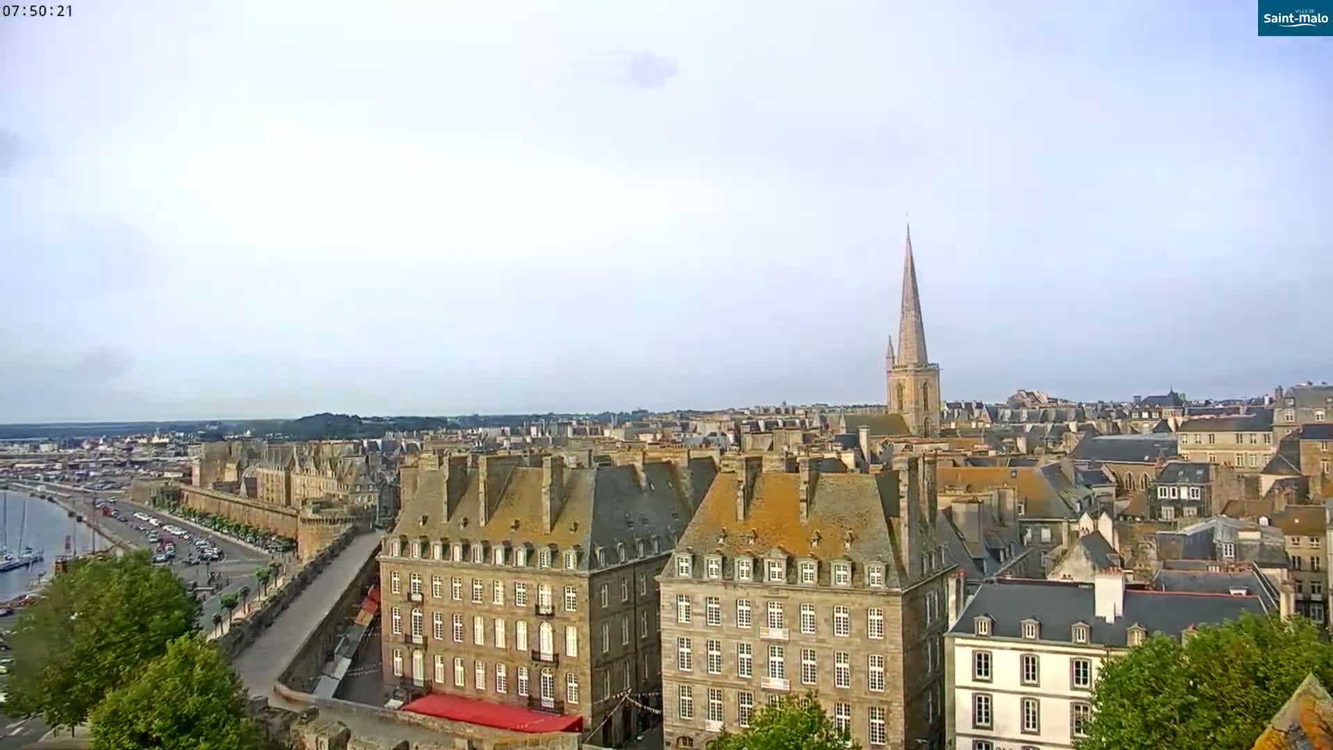 An elevated view of a coastal city on a partly cloudy day shows a collection of stone buildings with a prominent church steeple in the distance, a harbor with boats, and a walled section of the city.