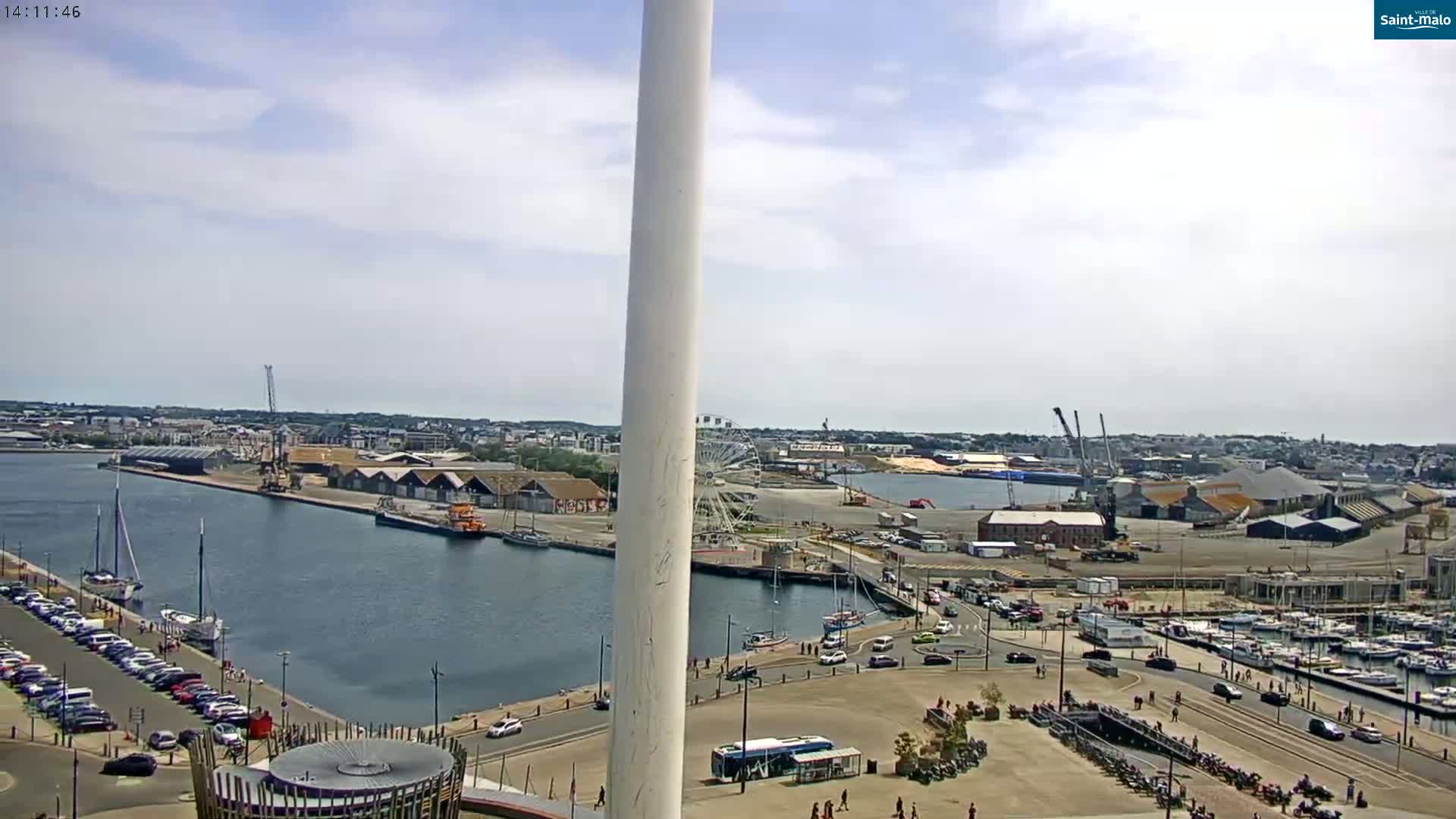 A partly sunny day overlooks a harbor with boats, a Ferris wheel, and industrial buildings, all viewed from a high vantage point near a tall, white structure.