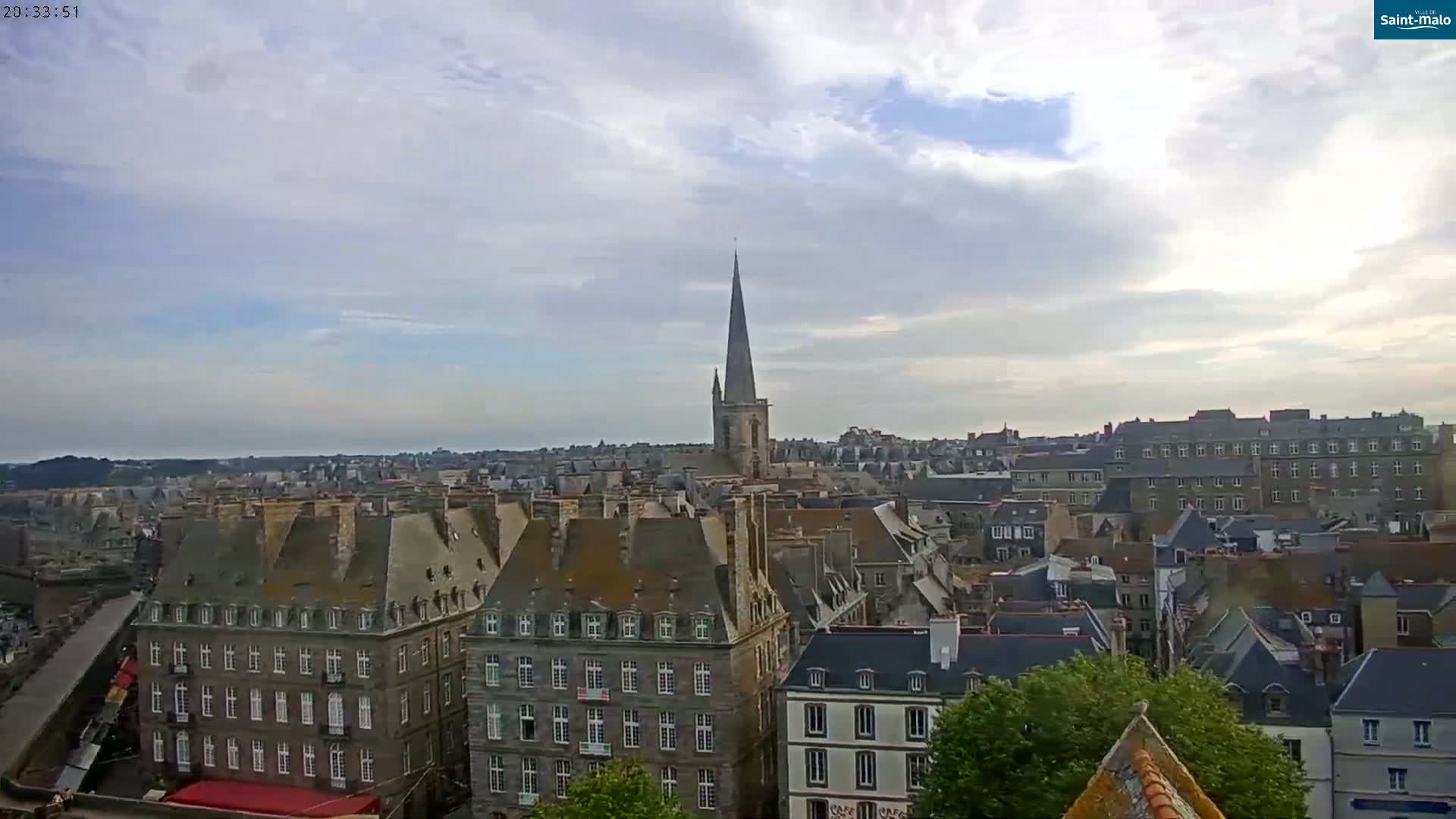 A partly cloudy sky overlooks a city of closely packed buildings with steeply pitched roofs and a tall church spire in the distance.