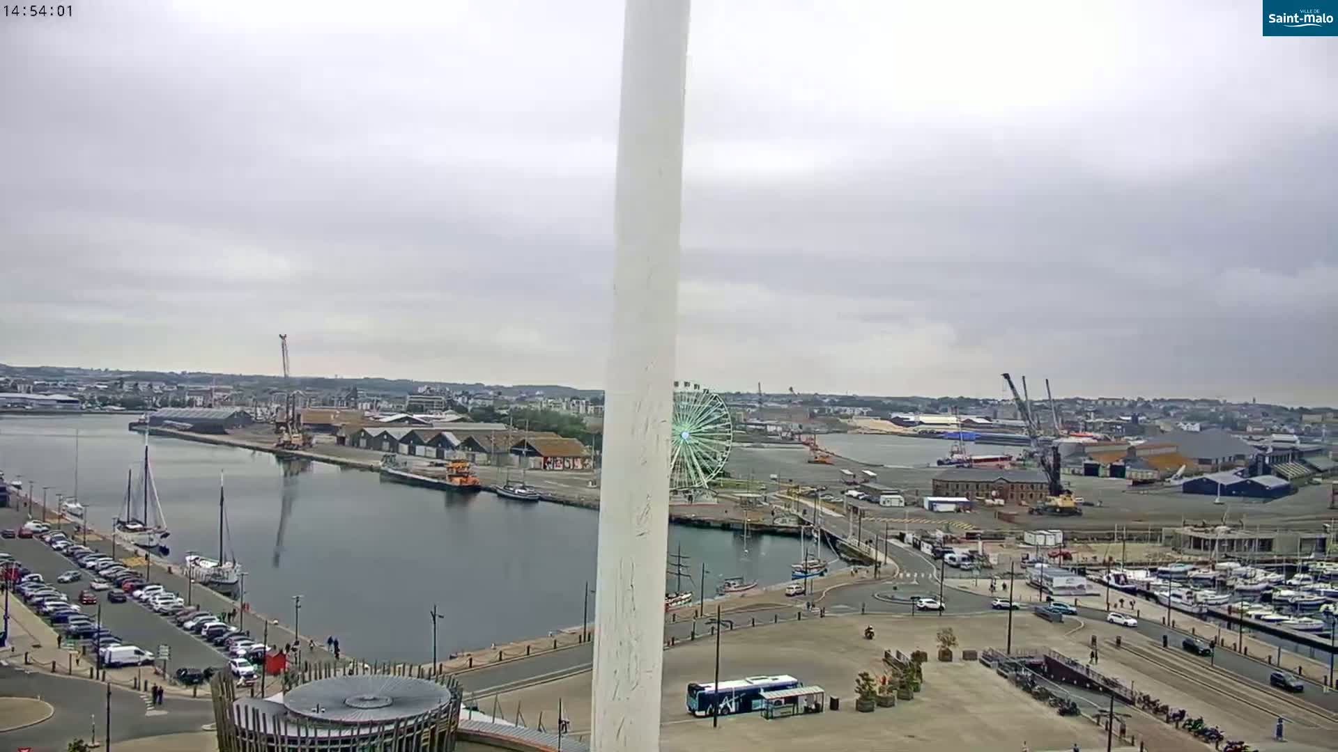 A harbor scene on an overcast day features a Ferris wheel, docked boats, and a tall, white structure dominating the foreground.