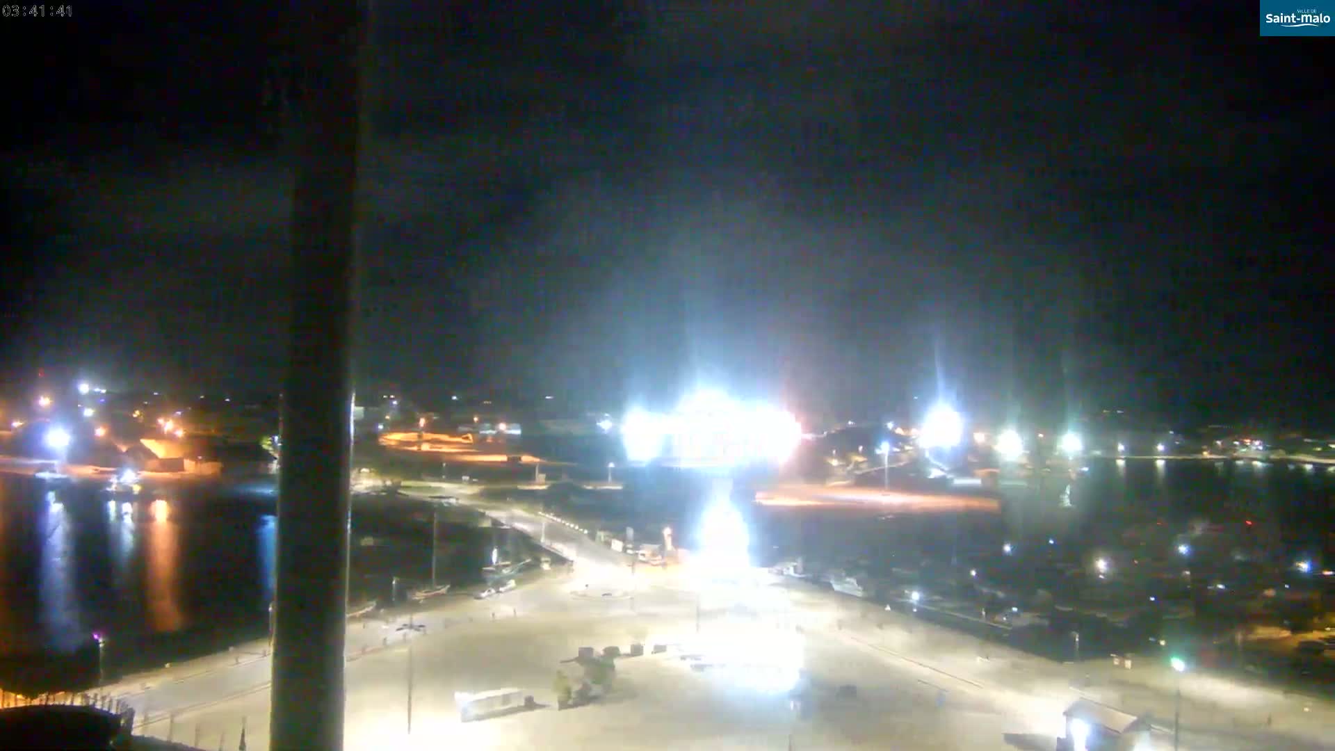 A nighttime, high-angle view of a harbor illuminated by artificial lights, with a bright light source in the center possibly from construction or a large vehicle.