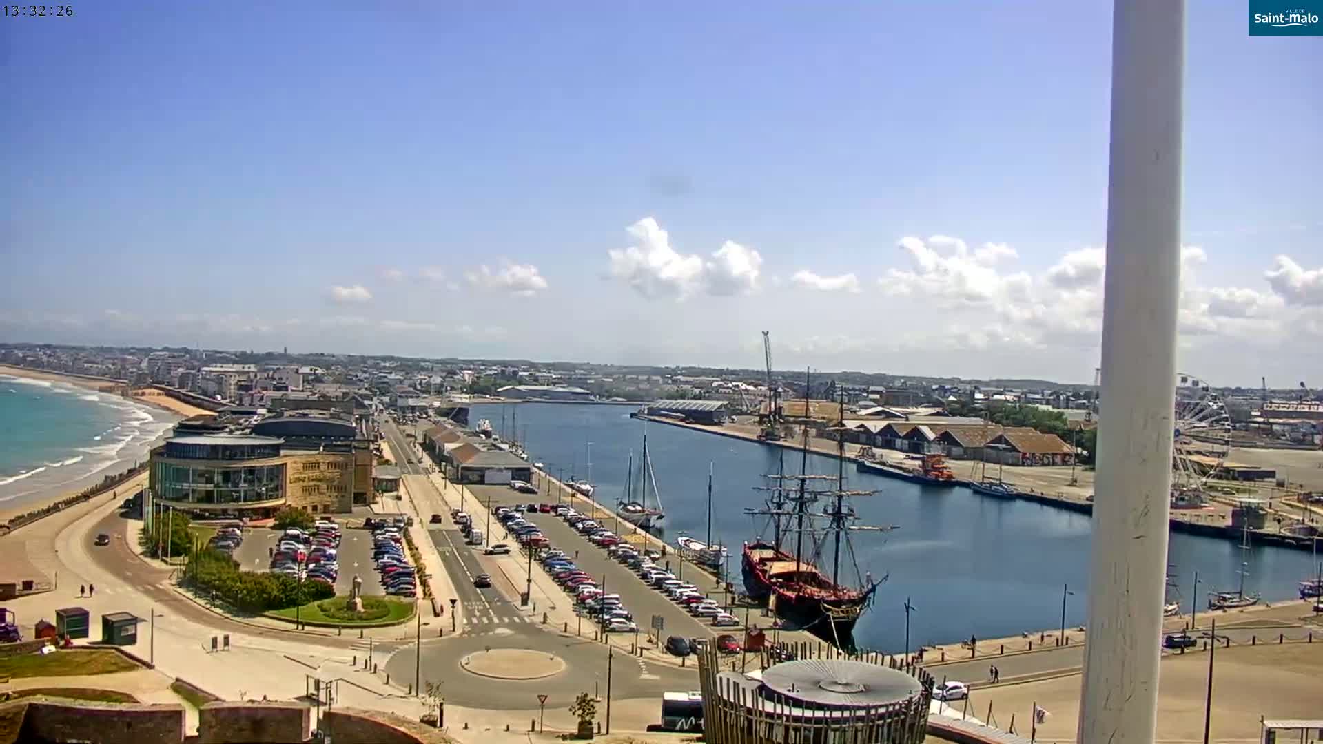 A high-angle view of a harbor on a sunny day with a partly cloudy sky, showing a sandy beach, docked ships, a large parking lot, and buildings along the waterfront.