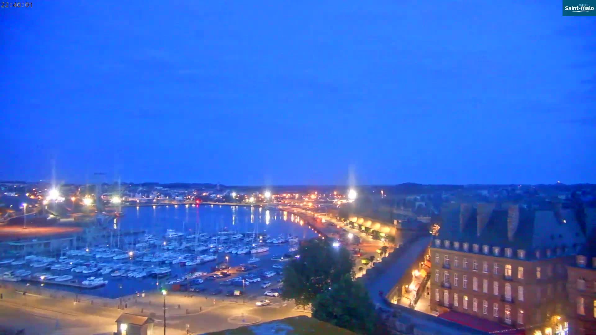 A nighttime, harbor view shows many boats docked in calm water, alongside a city with buildings lit up under a clear, deep blue twilight sky.