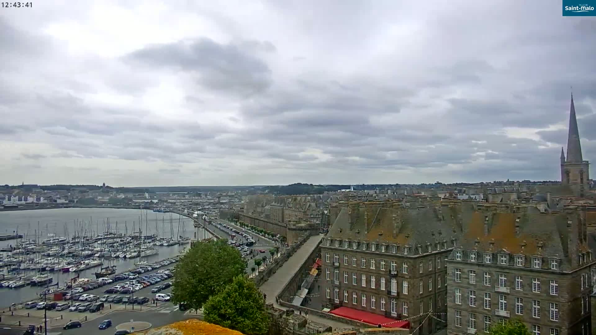 A harbor filled with sailboats is visible beneath a cloudy sky, next to a city with tall, grey buildings and a church spire.