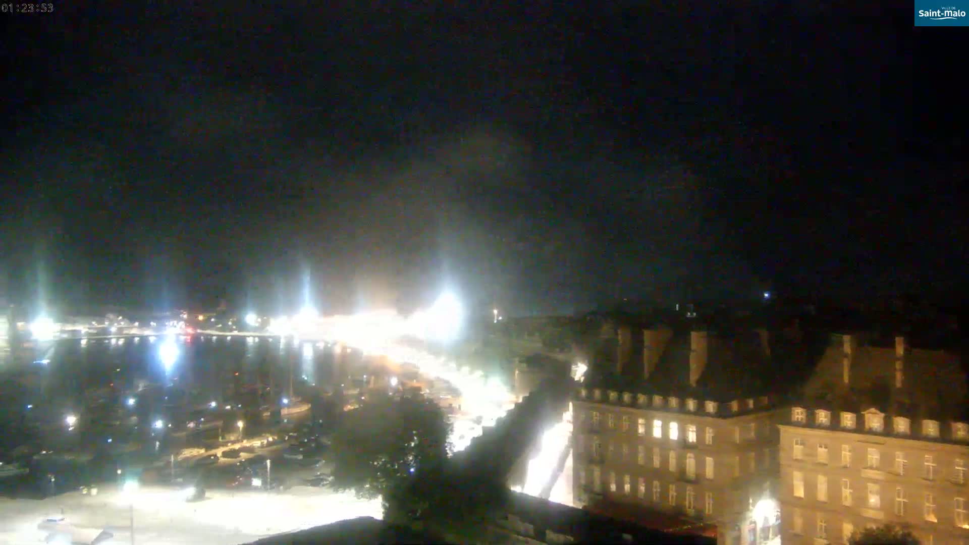A nighttime, high-angle view shows a harbor with boats, a city with illuminated buildings, and a dark sky.