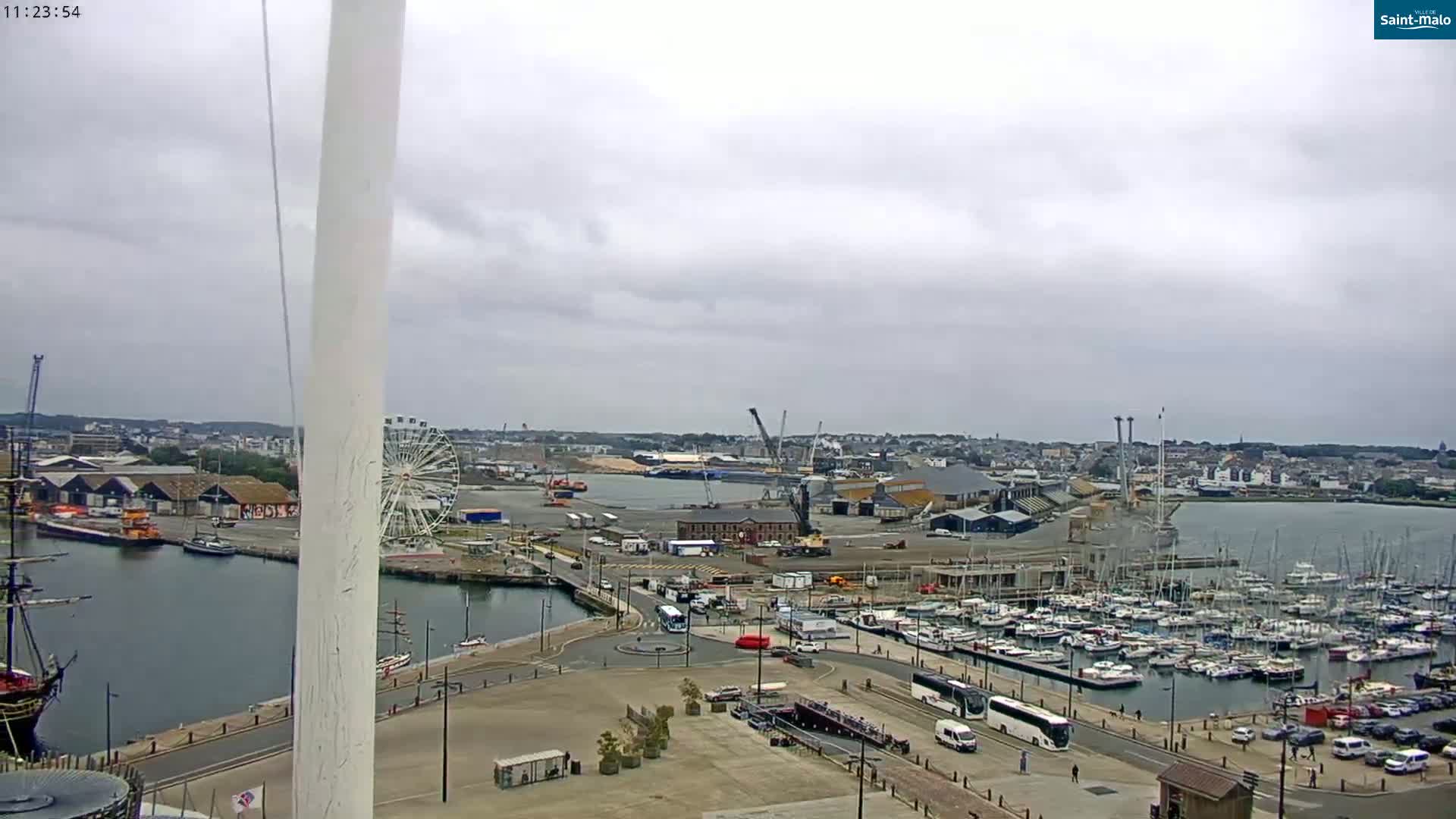 A harbor scene on an overcast day shows a marina filled with sailboats, a large ship, a Ferris wheel, and industrial areas, all viewed from a high vantage point.