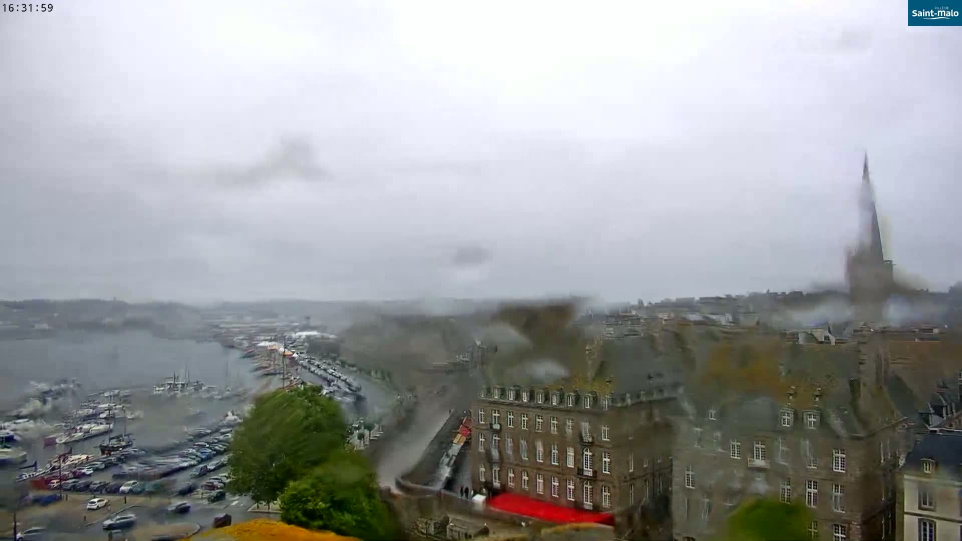 A rain-covered view of a harbor filled with boats and a city with stone buildings, one of which has a tall spire, under a gray, overcast sky.