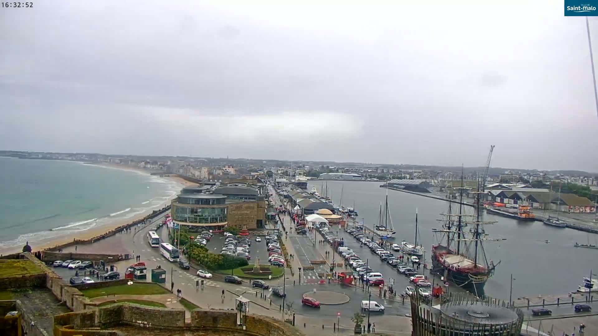 An overcast day reveals a coastal city scene with a harbor filled with boats, including a large sailing ship, and a sandy beach with a curving road lined with parked cars.