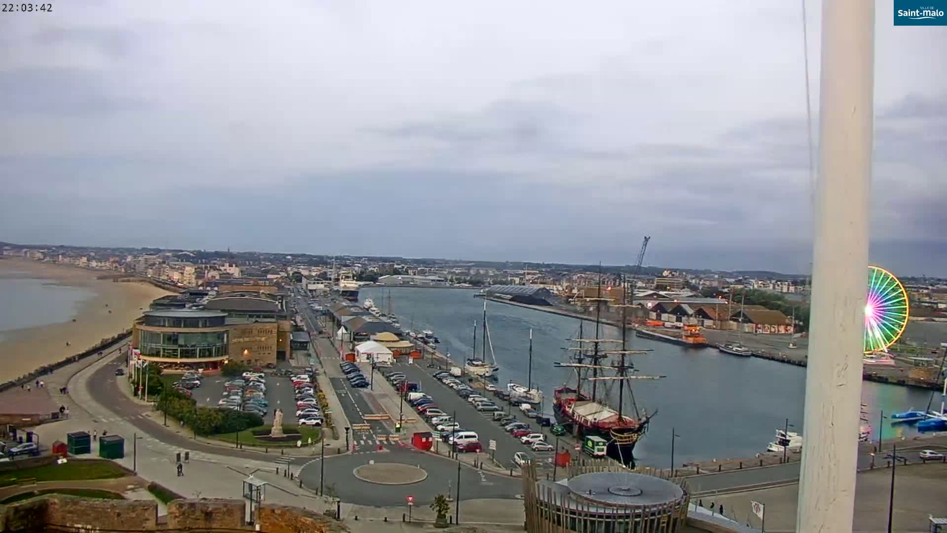 An elevated view of a harbor town on an overcast day shows a sandy beach, a large parking lot, buildings, a tall ship docked in the harbor, and a Ferris wheel.
