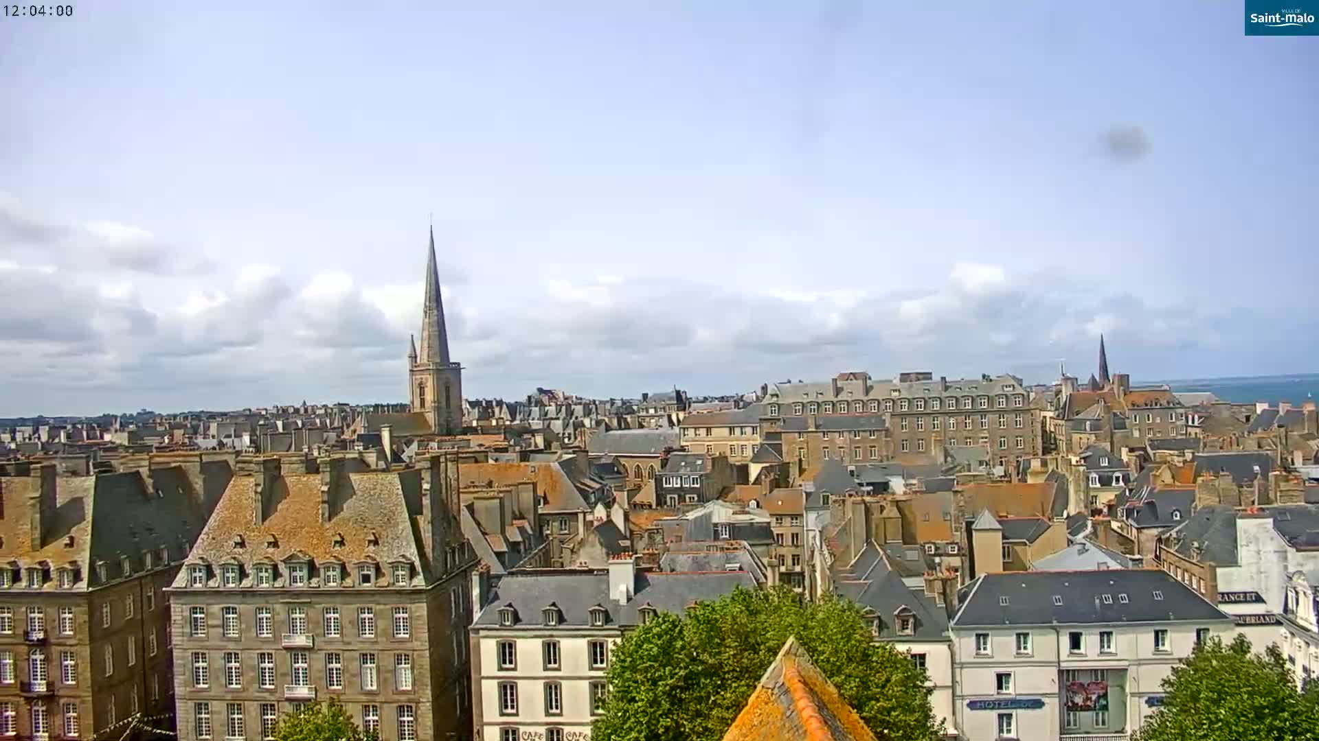 A partly cloudy day reveals a panoramic view of a European city with numerous slate-roofed buildings, a prominent church steeple, and a glimpse of the ocean in the distance.