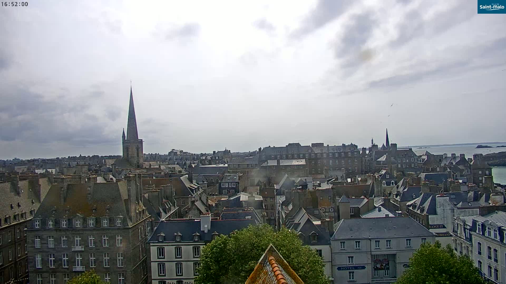 An overcast day reveals a sprawling cityscape of grey-roofed buildings, punctuated by a tall church spire and overlooking a calm body of water.