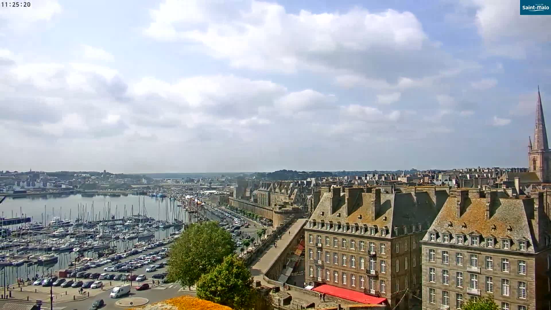 A partly cloudy day overlooks a harbor filled with sailboats and a city of buildings with steeply pitched roofs.