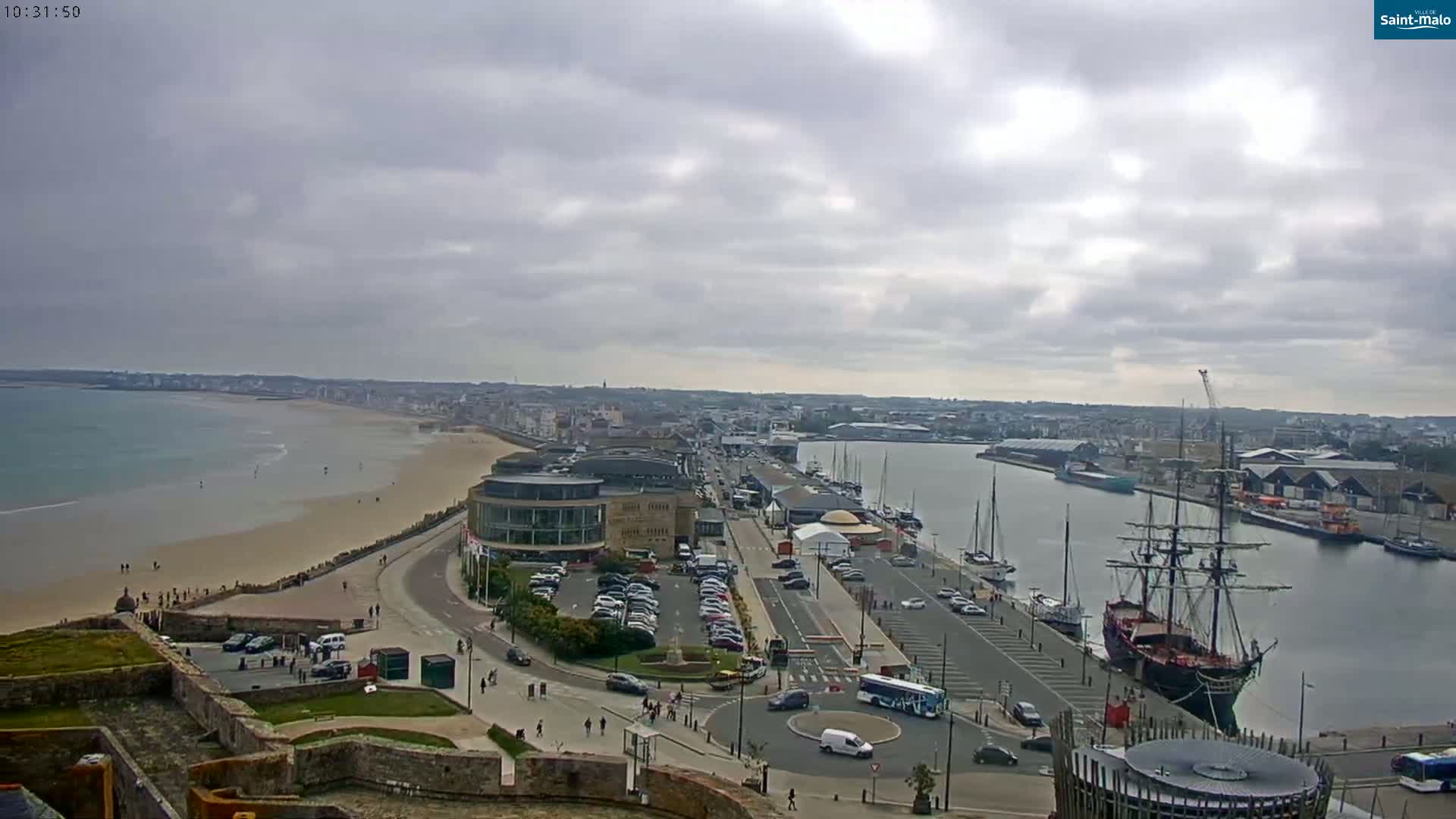 An overcast day reveals a coastal city with a sandy beach, harbor filled with boats, including a large sailing ship, and a parking lot next to buildings.
