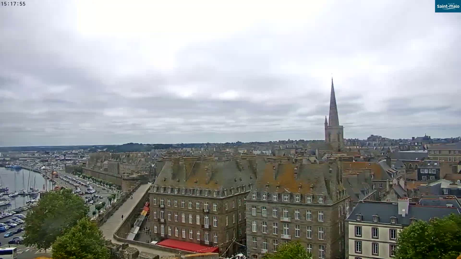 A high-angle, long shot of a coastal city on an overcast day, showing a harbor filled with boats, a city wall, and many buildings with steeply pitched roofs, including a tall church spire.
