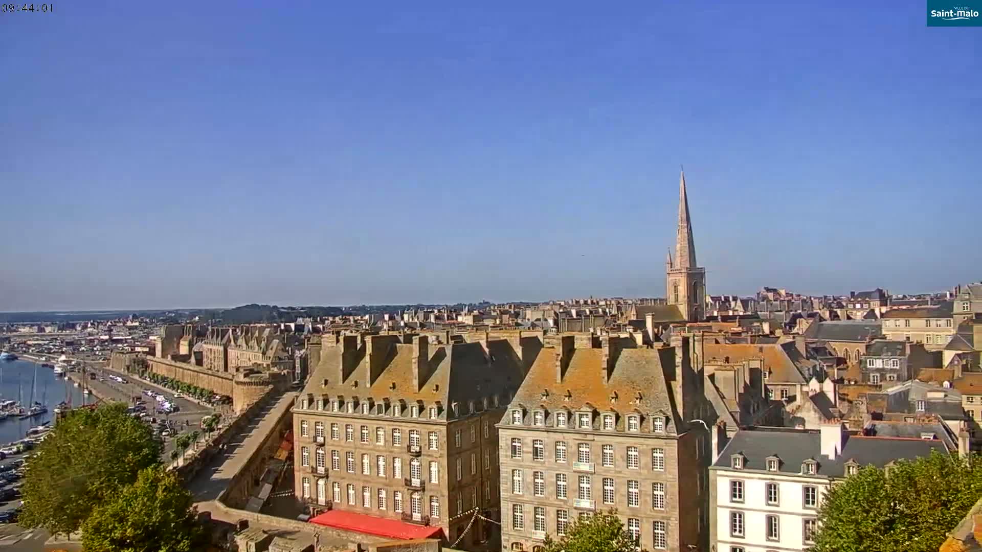 A high-angle, long shot on a clear, sunny day shows a coastal city with a harbor, stone buildings, and a tall church steeple in the distance.