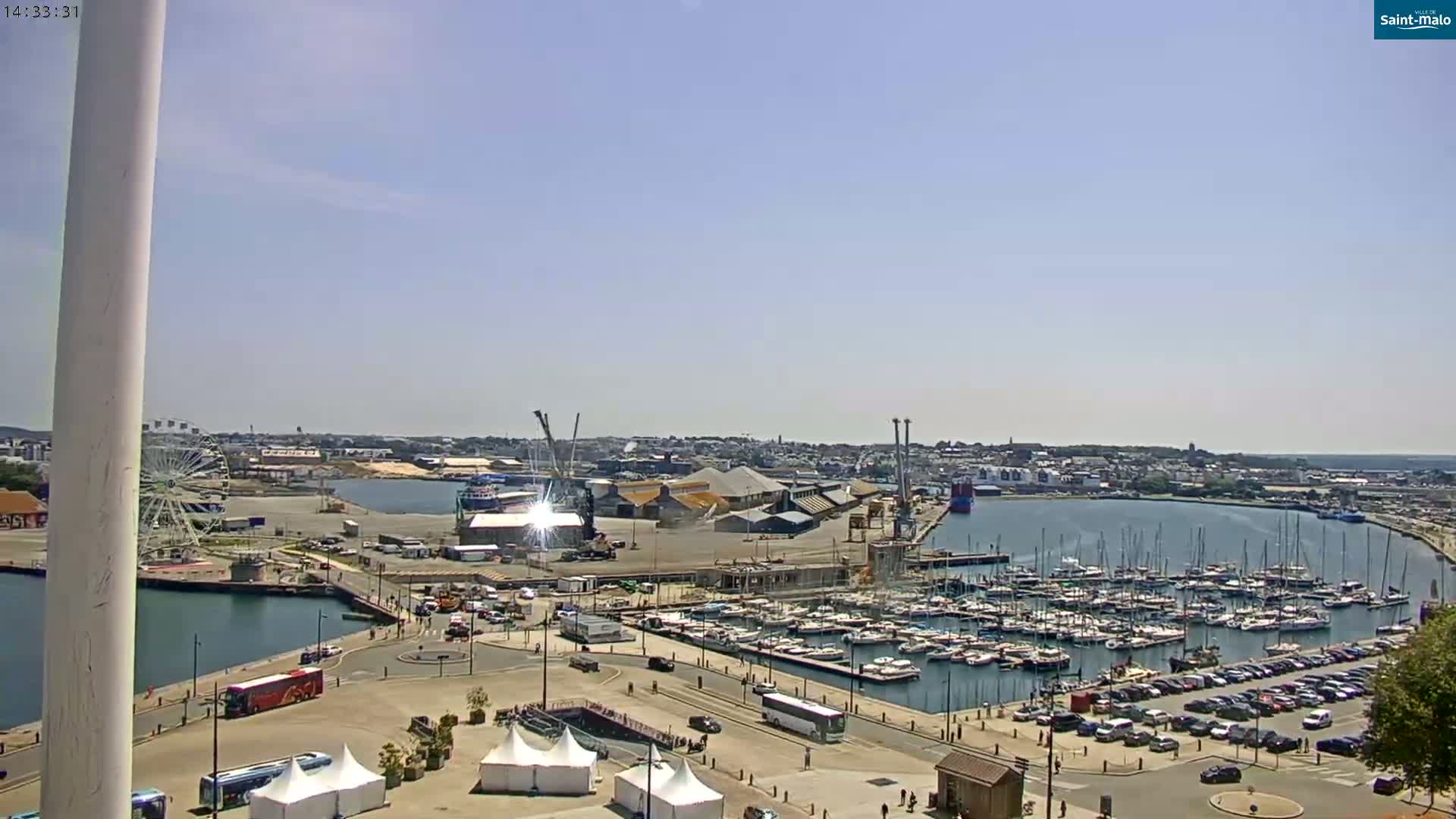 A sunny day reveals a harbor filled with numerous boats and yachts, adjacent to a city and a Ferris wheel, with a large parking lot nearby.