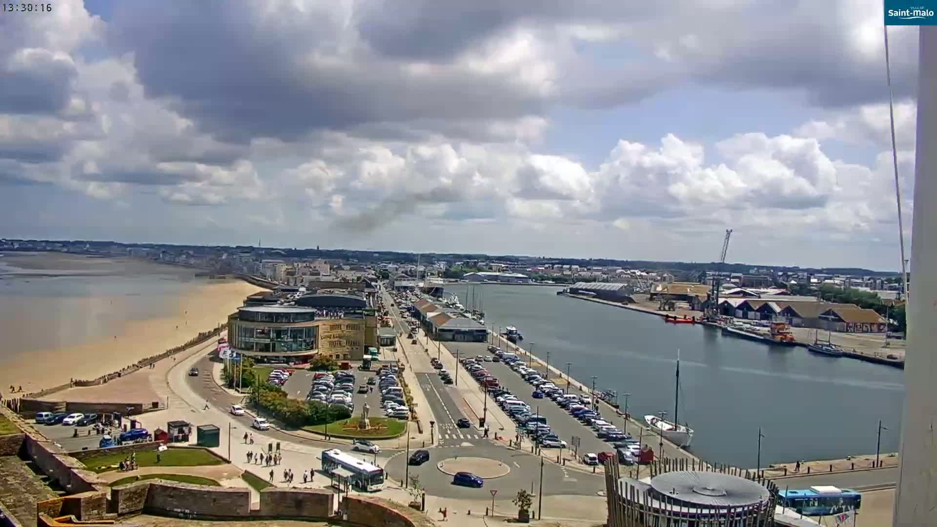 A coastal city with a harbor, sandy beach, and buildings is visible under a partly cloudy sky.