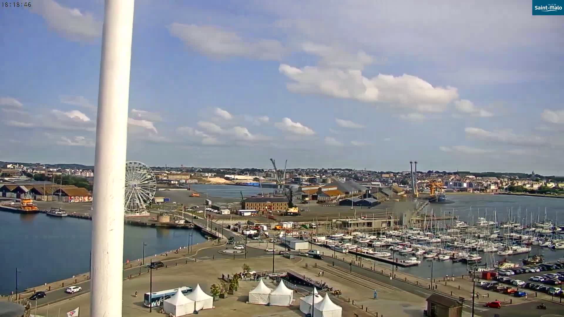 A partly cloudy day overlooks a harbor filled with sailboats and a Ferris wheel, with a city in the background.