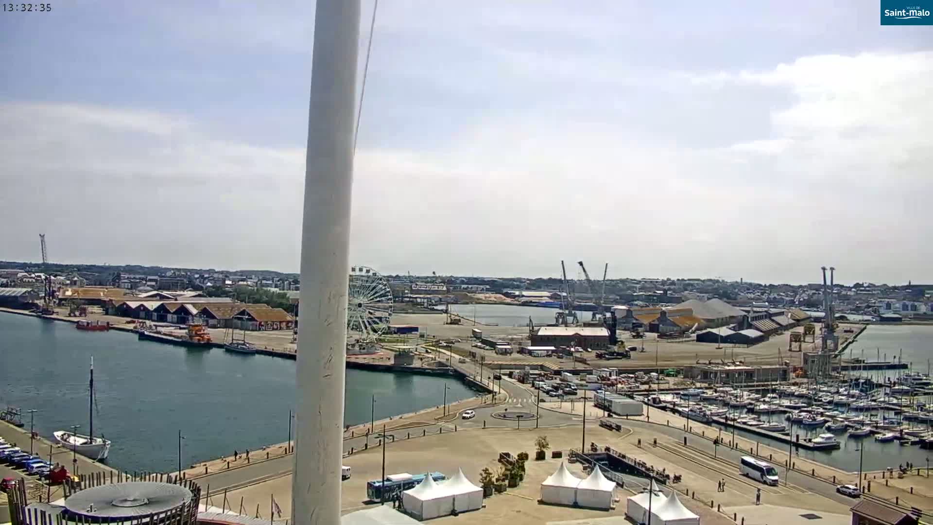 An elevated view of a harbor on a partly sunny day shows a marina filled with boats, a Ferris wheel, industrial buildings, and a waterfront area with several white tents.