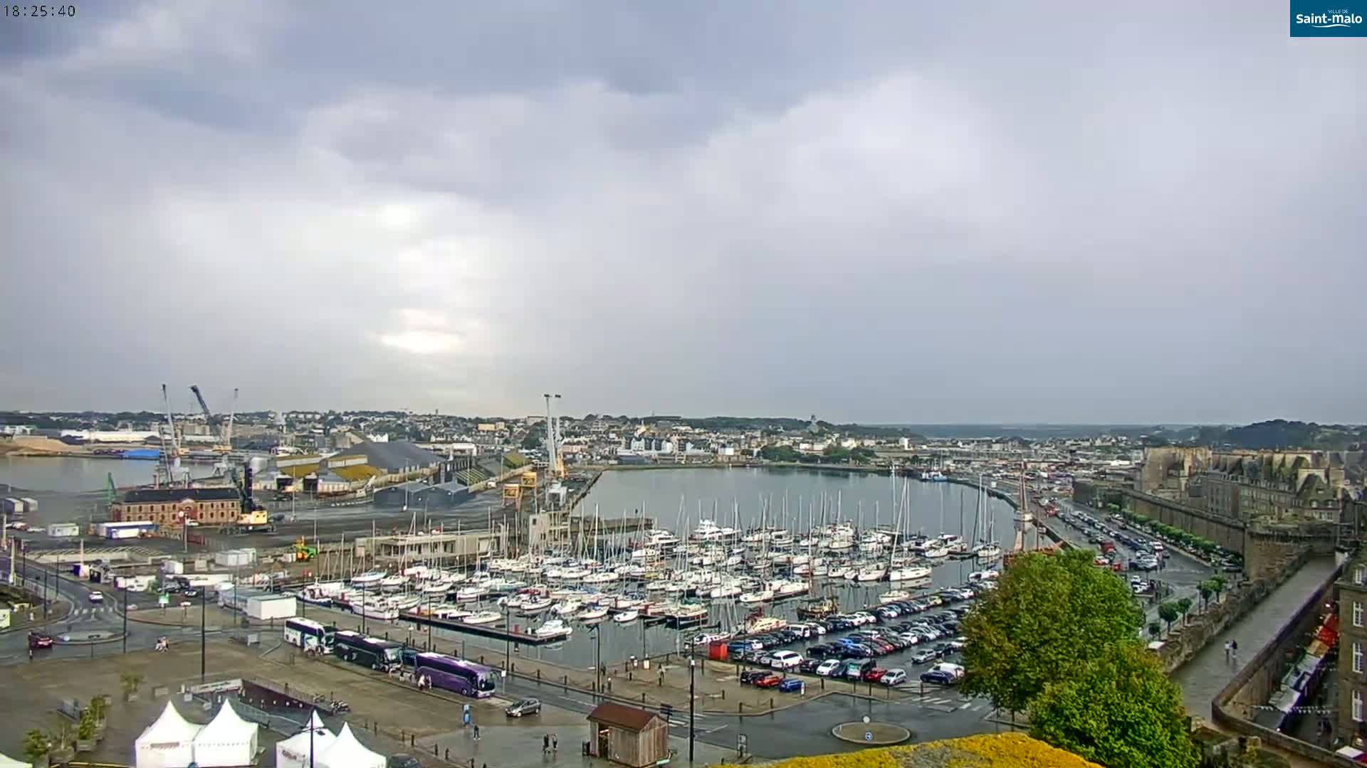A harbor filled with sailboats and yachts is shown under a mostly cloudy sky, with a city and a walled area visible in the background.